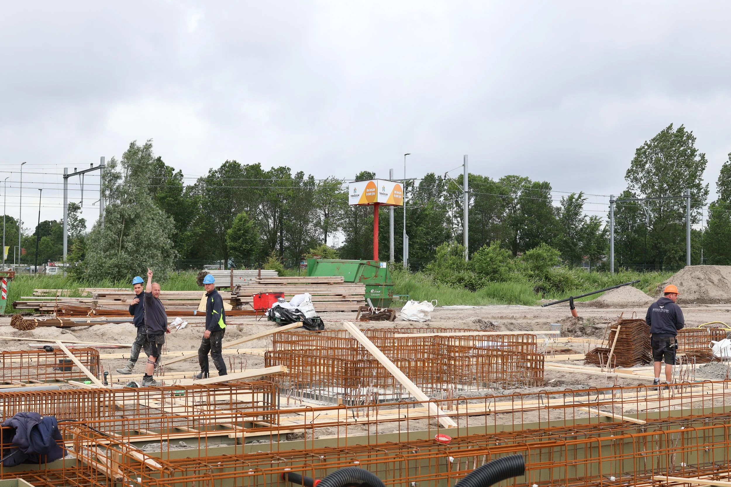 Constructieplaats met betonfunderingen en bouwers in veiligheidshelmen, omgeven door bouwmaterialen en apparatuur, onder een bewolkte lucht.