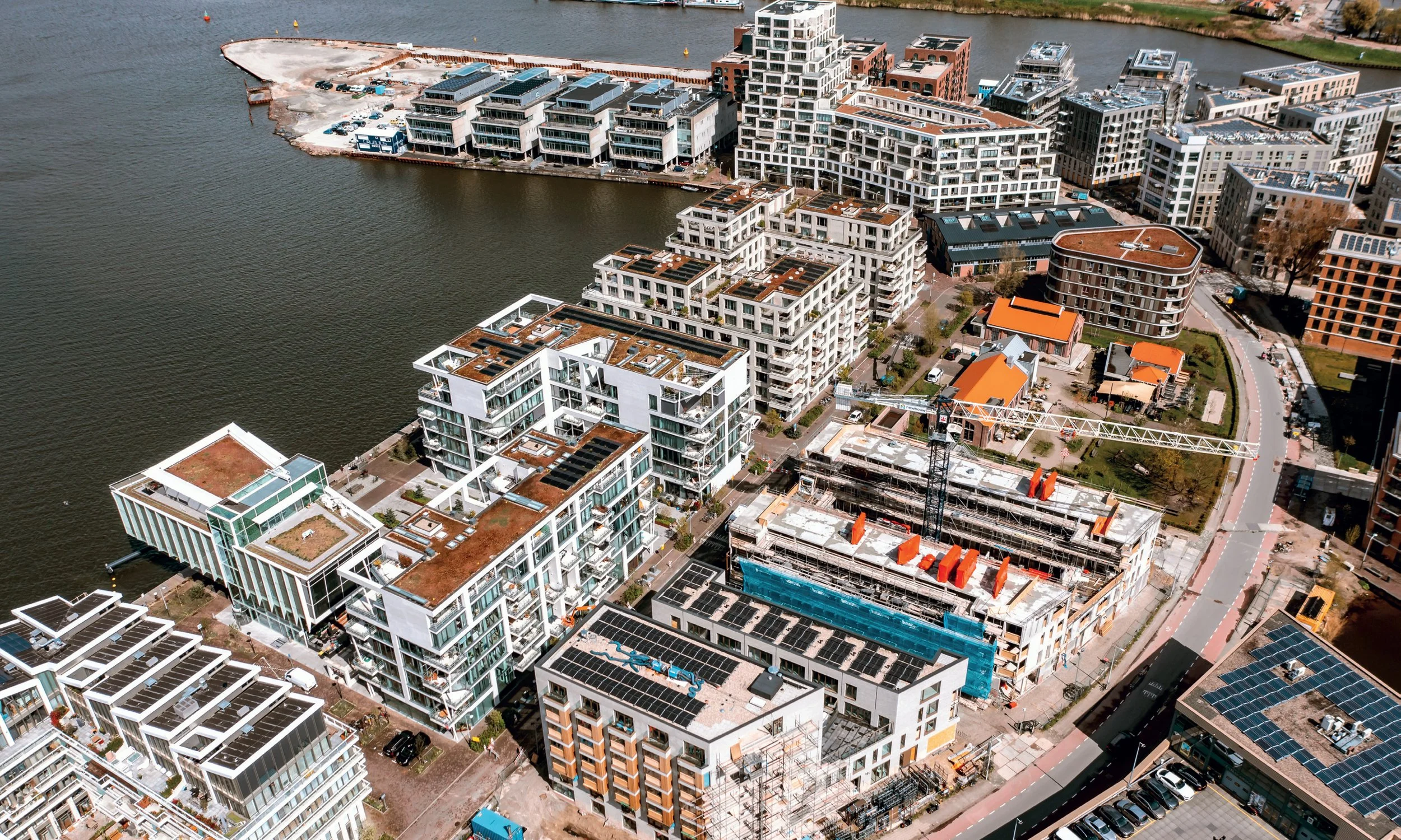 Luchtfoto van een stedelijk gebied met moderne appartementsgebouwen langs een rivier, met bouwplaatsen en parkeergelegenheid, en gedeeltelijk blauwe hemel.