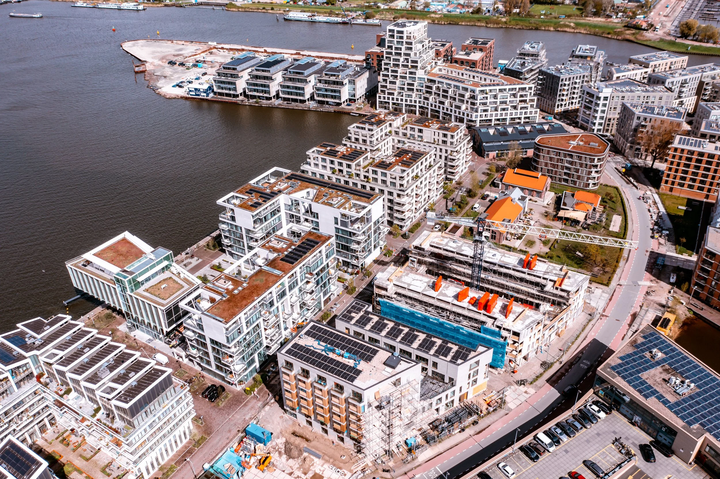 Uitzicht op moderne appartementsgebouwen langs een rivier met bootjes, een onder constructie zijnde gebouw met een bouwkraan, en parkeergelegenheid in de stad.