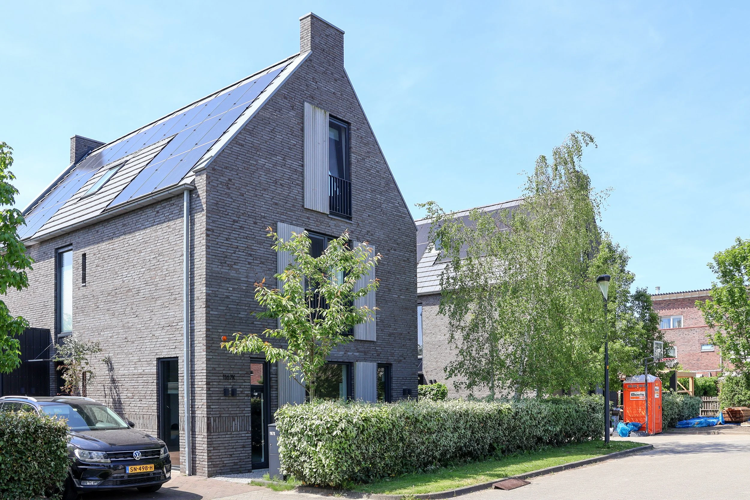 Moderne woning met grote ramen, een dak met zonnepanelen en een kleine balkon. Bomen en planten rondom, geparkeerde auto's op de straat.