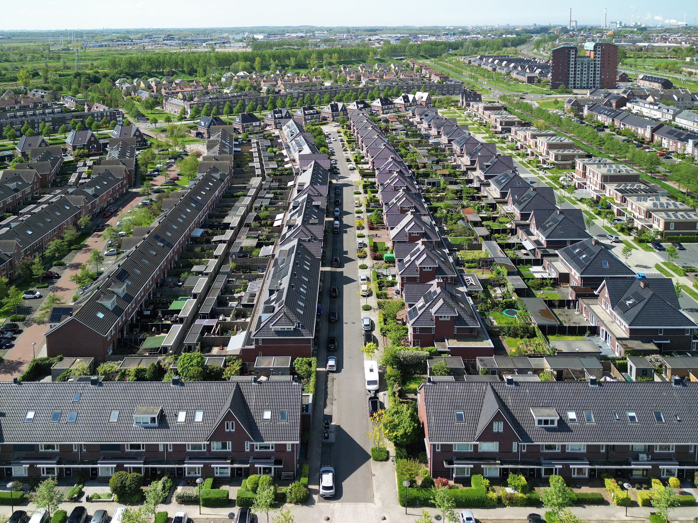 Een bird's-eye view van een woonwijk met rijtjeshuizen, groene tuinen en bomen, met in de achtergrond een industriegebied en een open landschap.