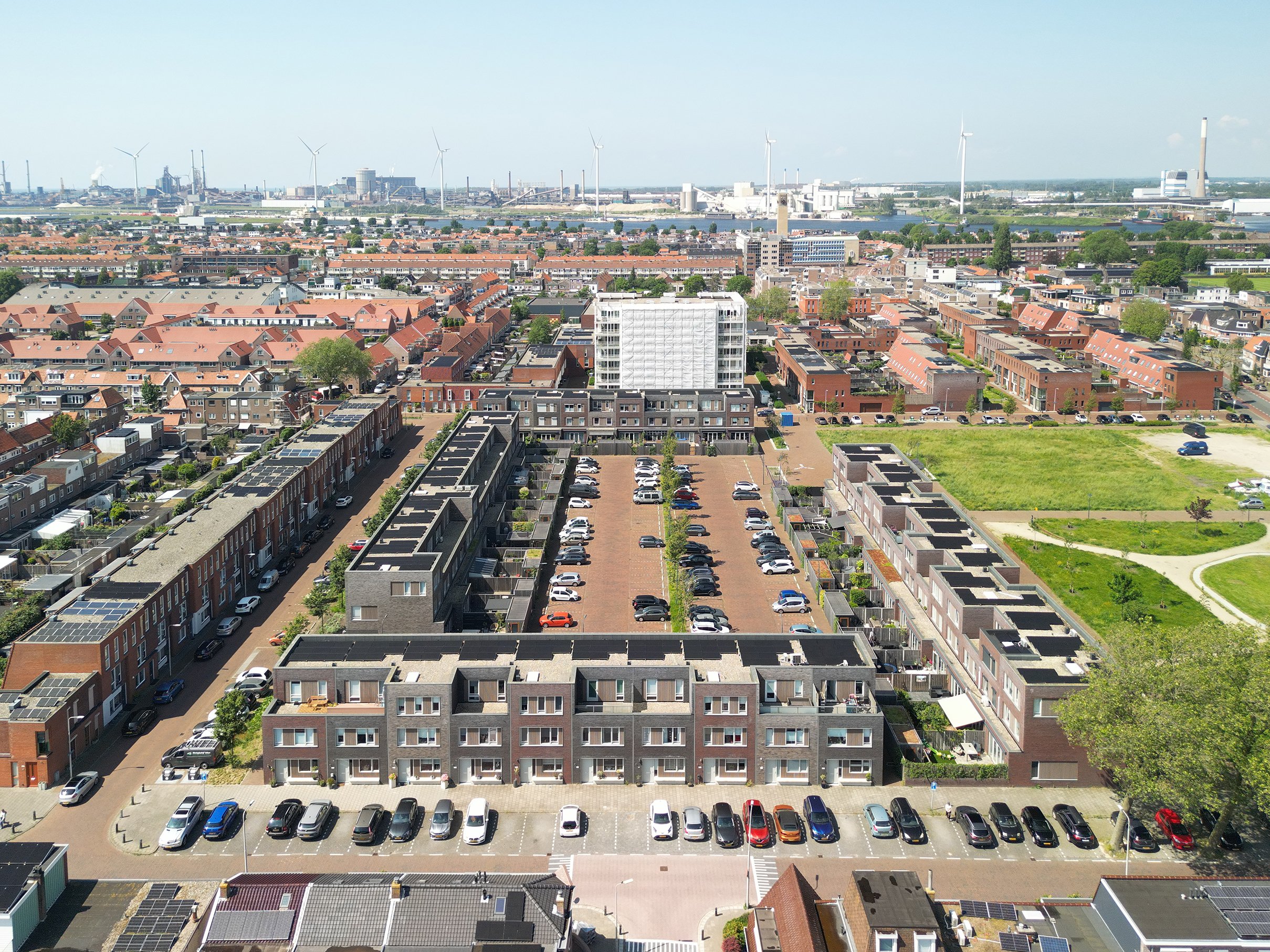 Uitzicht over een woonwijk met rijen rijtjeshuizen en parkeerplaatsen, met een industriegebied en windturbines op de achtergrond onder een blauwe hemel.