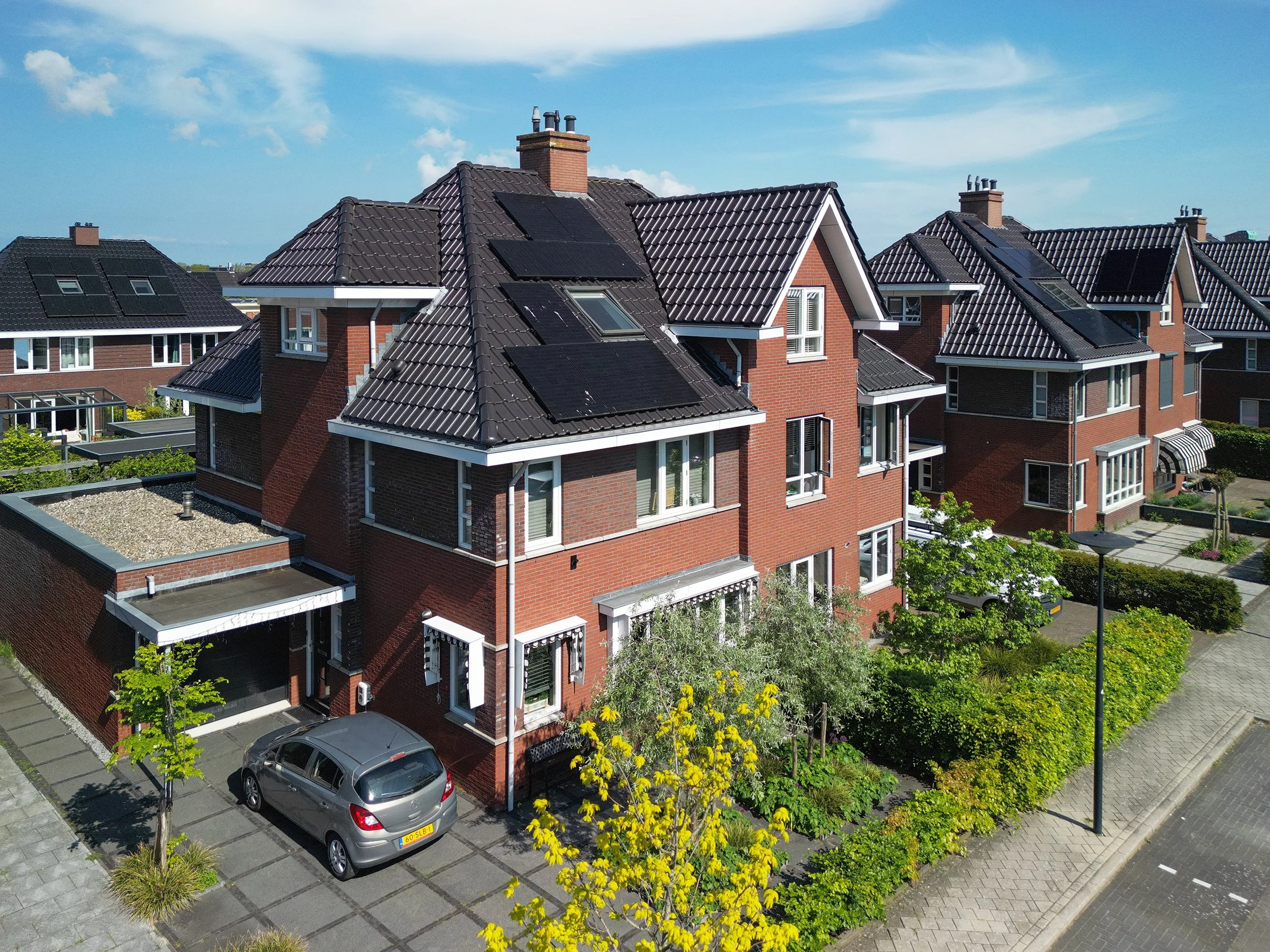 Twee rijtjeshuizen met rode bakstenen muren en zwarte dakpannen, met zonnepanelen en schoorstenen, onder een blauwe lucht met enkele witte wolken.
