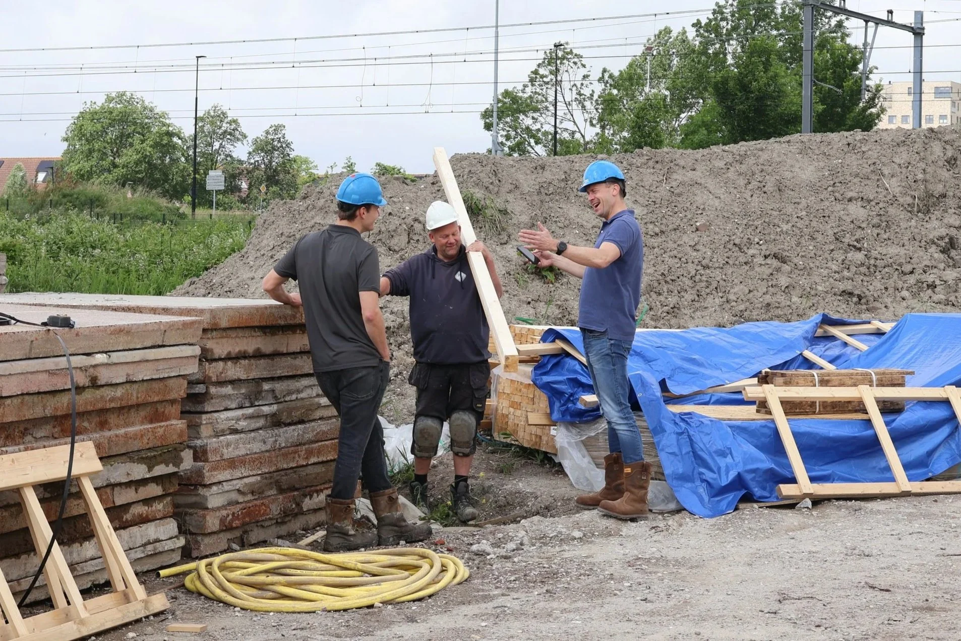 Drie bouwvakkers met helmen lachen en praten op een bouwplaats, omgeven door bouwmaterialen zoals stenen, hout en elektrische kabels.