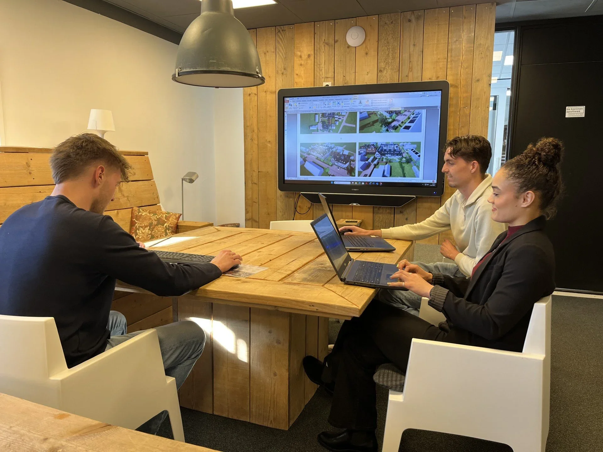 Vijf mensen werken samen aan een project in een vergaderruimte met houten wanden en een groot beeldscherm met ontwerpplannen.