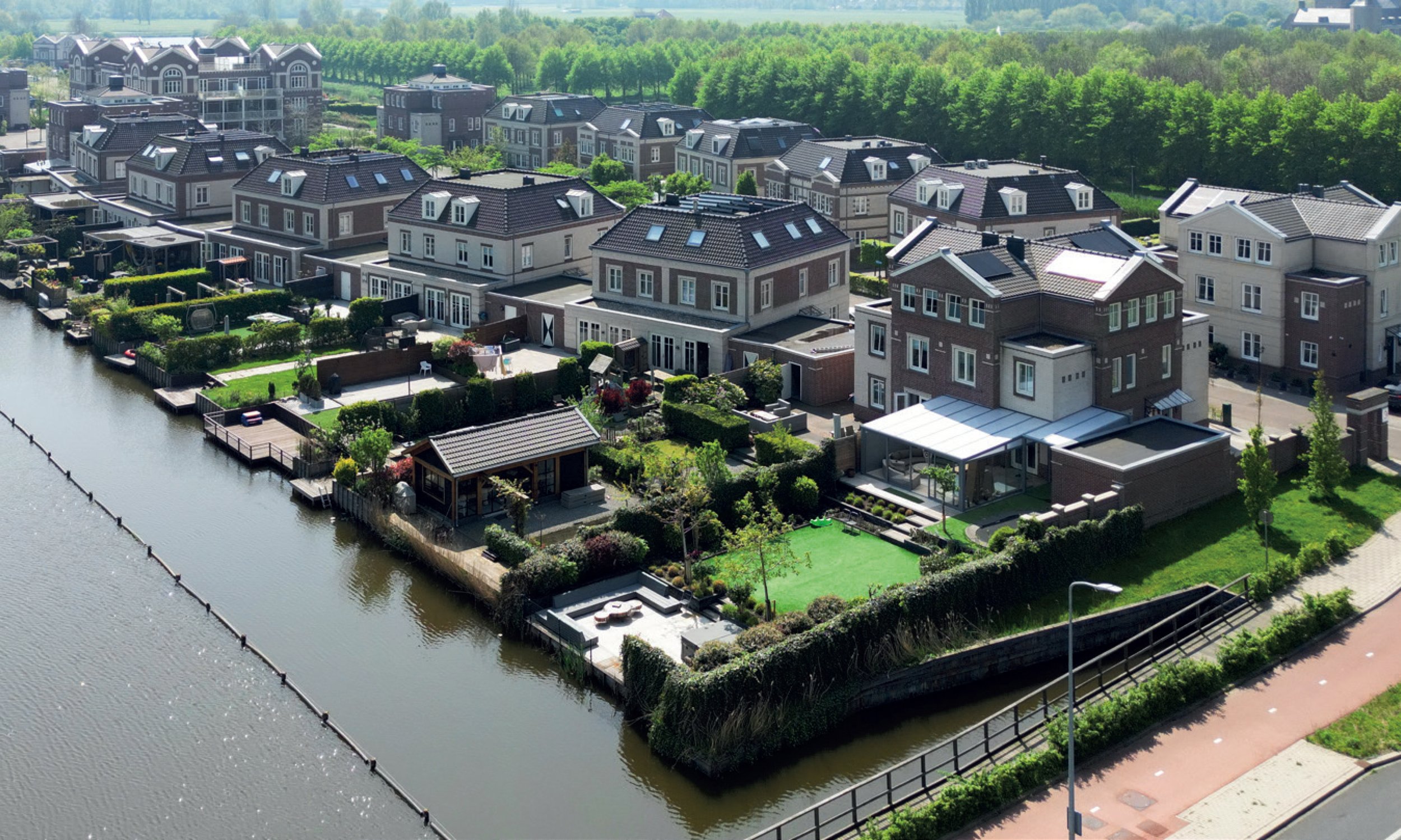 Een overzicht van een woonwijk met moderne huizen langs een waterkant, omringd door groene bomen en met een fietspad ernaast.