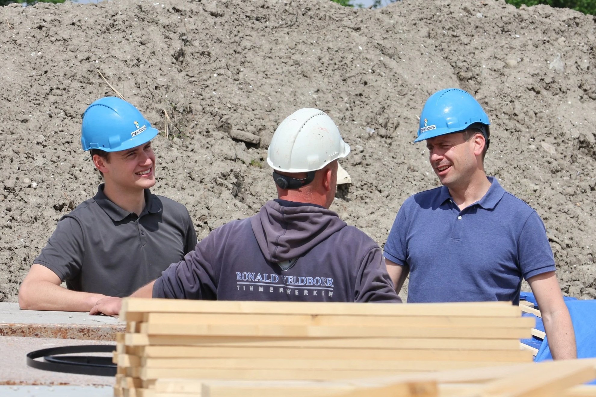Drie mannen met helmen die lachen en praten op een bouwplaats.