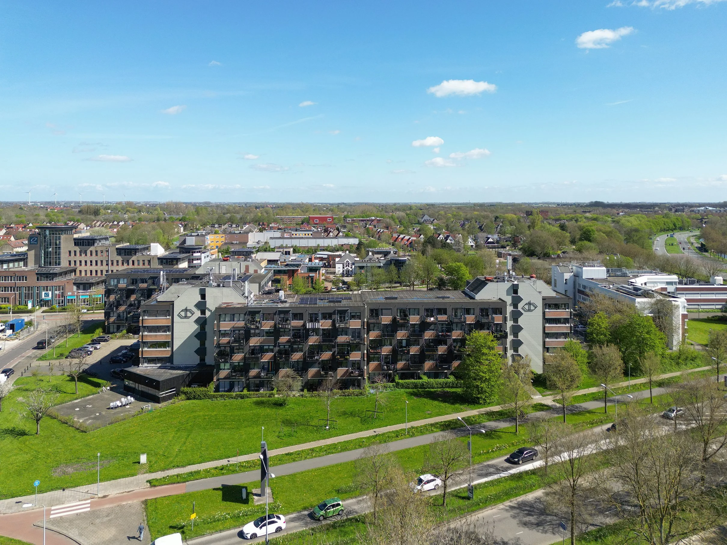 Een stadsuitzicht met moderne appartementen, groene parken, wegen met auto’s, en een heldere blauwe lucht met een paar witte wolken.