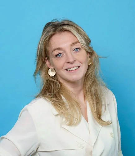 Een vrouw met blond haar en blauwe ogen, glimlachend, tegen een blauwe achtergrond.