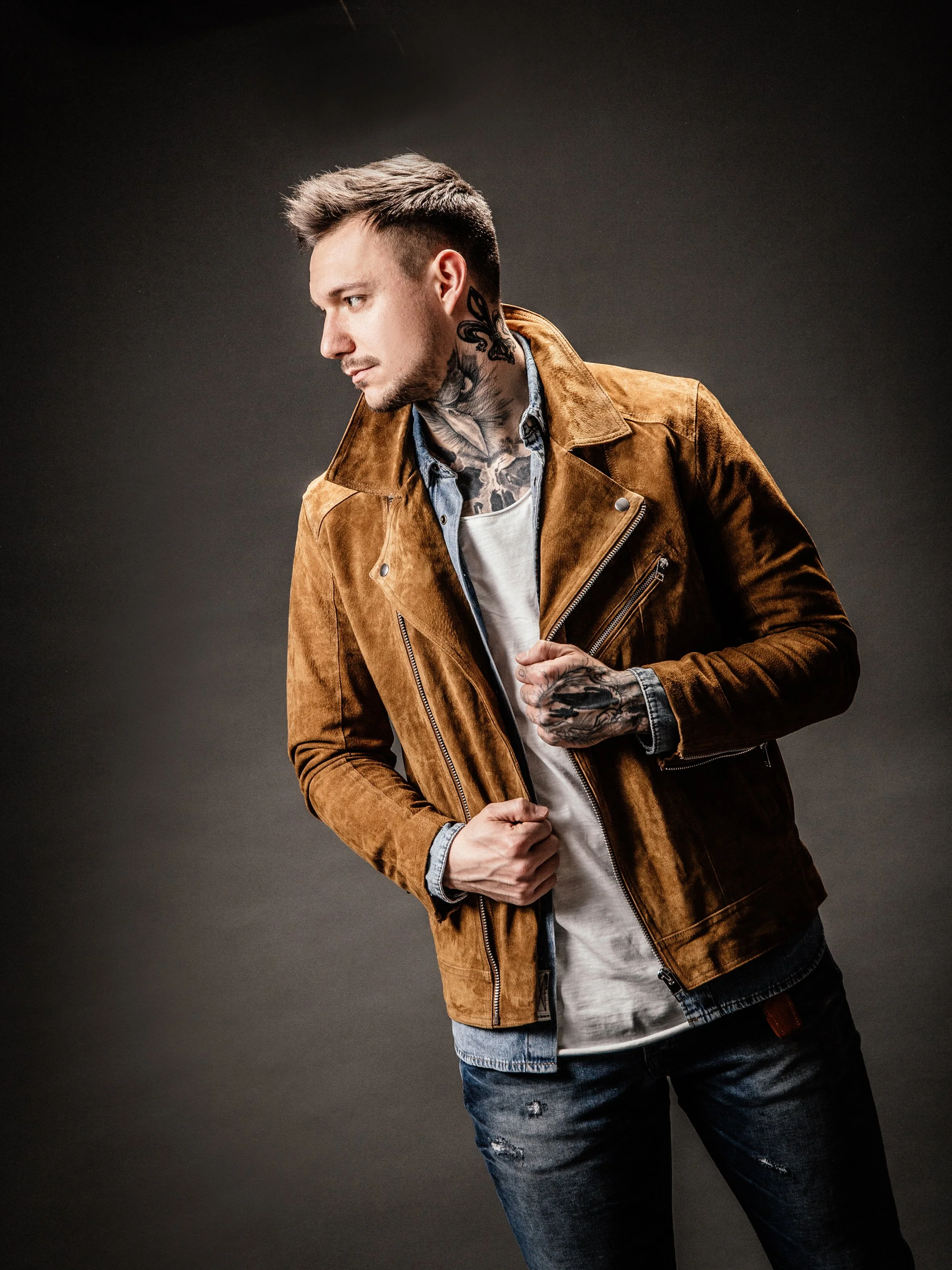 Junger Mann mit Tattoos in reflexiver Pose, trägt braune Lederjacke, graues T-Shirt und Jeans, gegen dunklen Hintergrund.