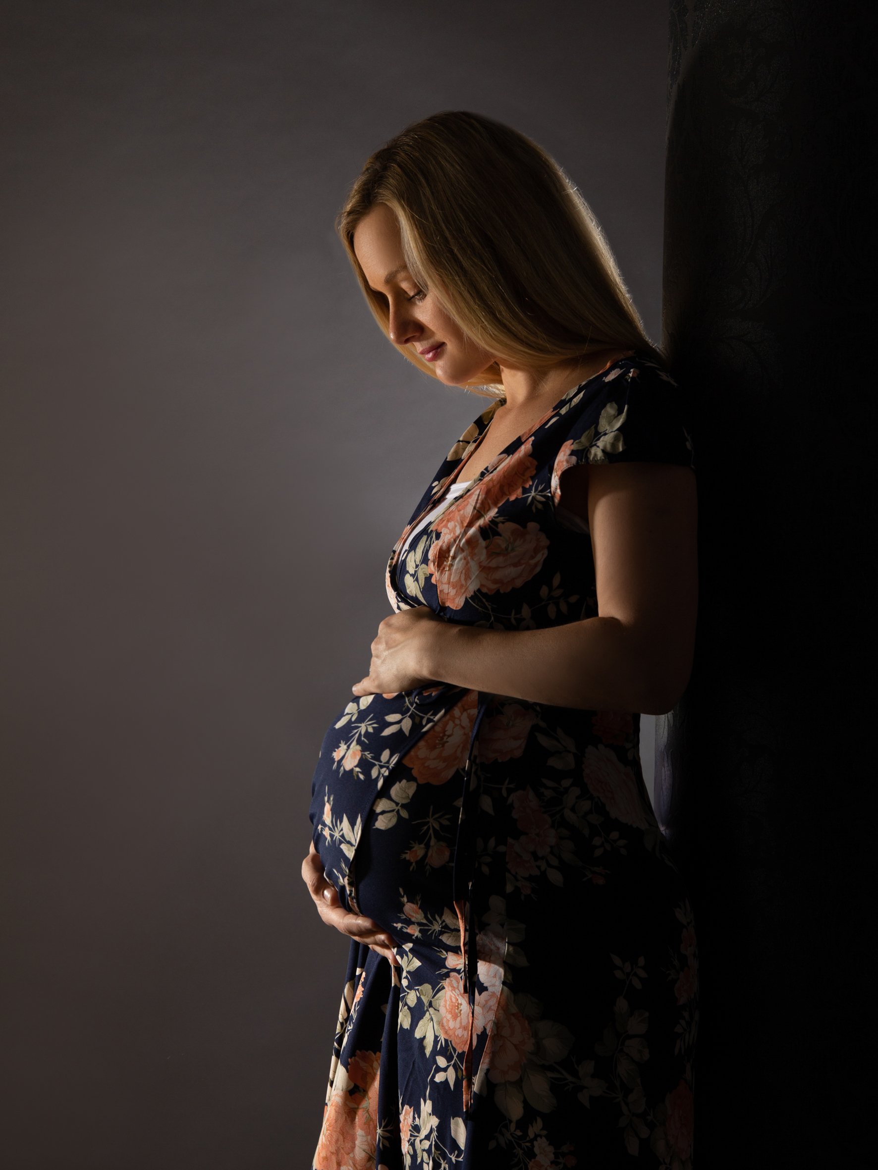 Eine schwangere Frau mit langem Haar, die an einer dunklen Wand lehnt, trägt ein florales Kleid und hält ihr Babybäuchlein.