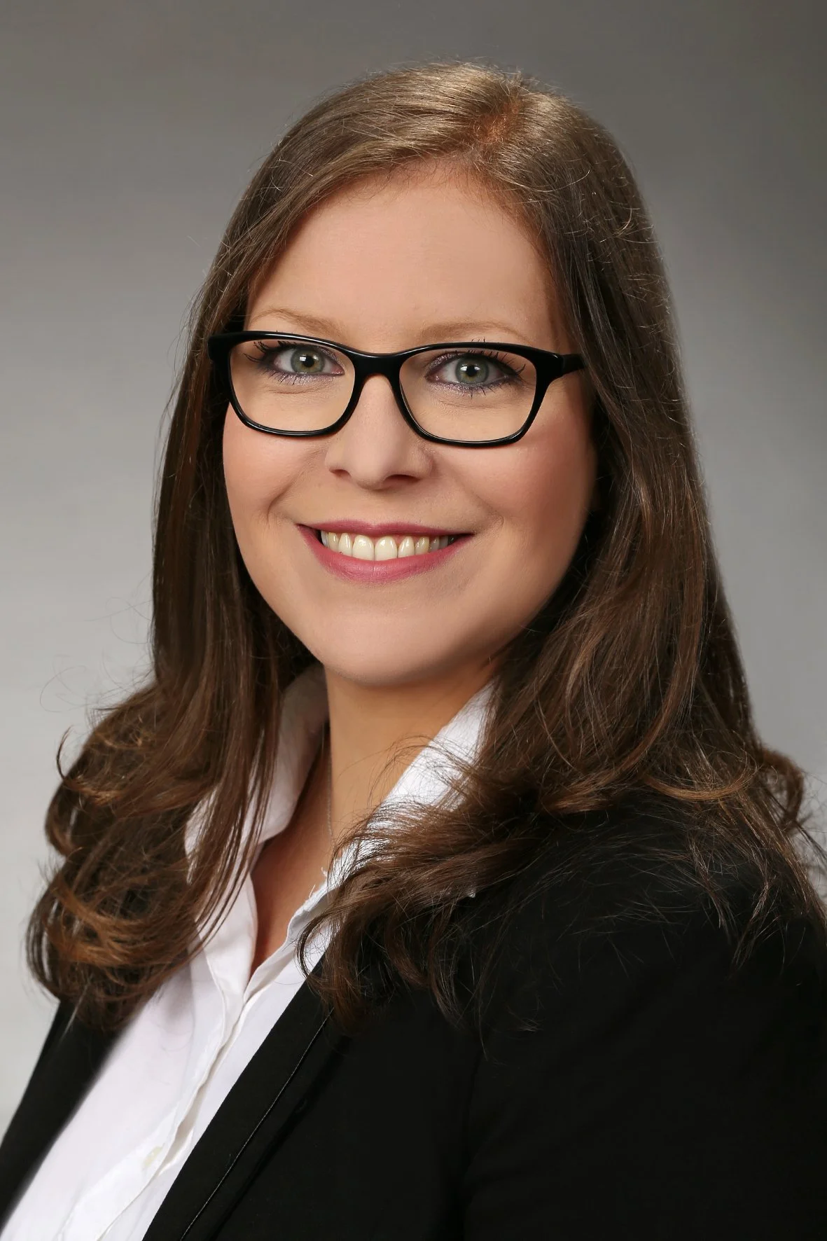 Porträt einer lächelnden Frau mit braunen, gewellten Haaren, schwarzen Brillen, weißem Hemd und schwarzer Jacke vor grauem Hintergrund.