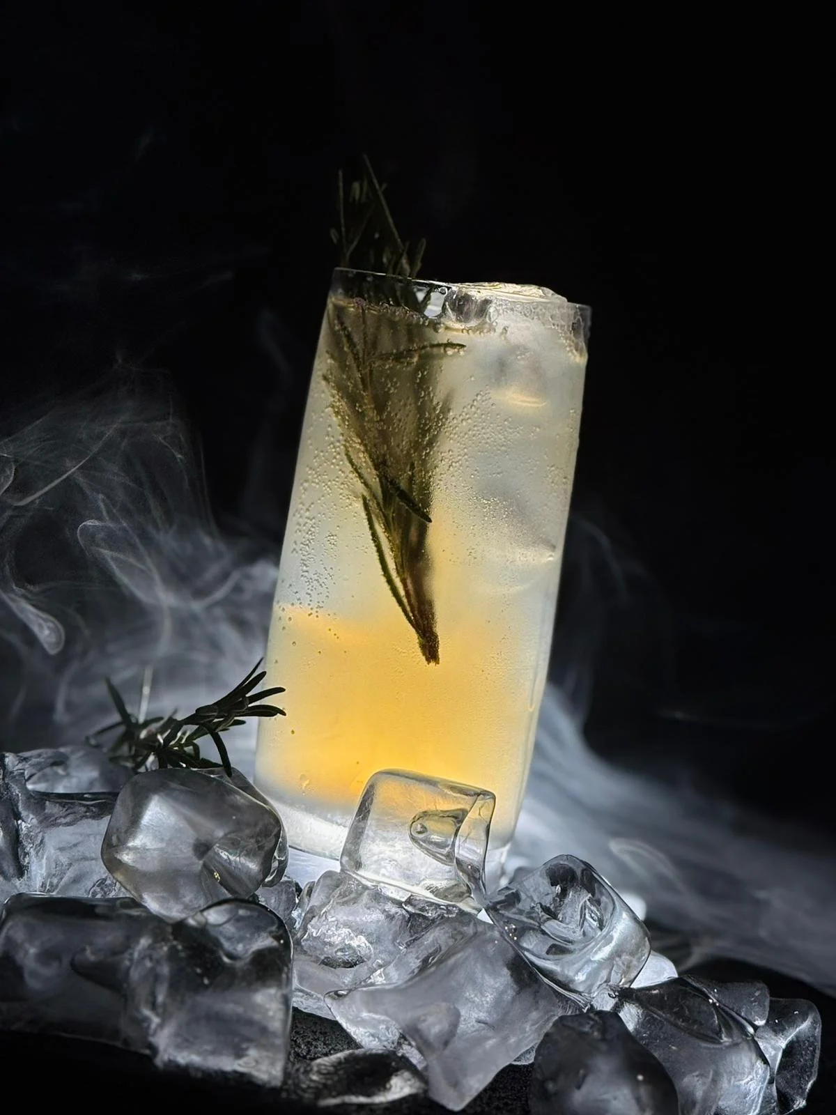 Ein hohes Glas mit einem sprudelnden Getränk, darin befinden sich Eiswürfel und ein Rosmarinzweig. Das Glas steht auf Eiswürfeln und einem schwarzen Hintergrund, Rauch- oder Nebeleffekt im Hintergrund.