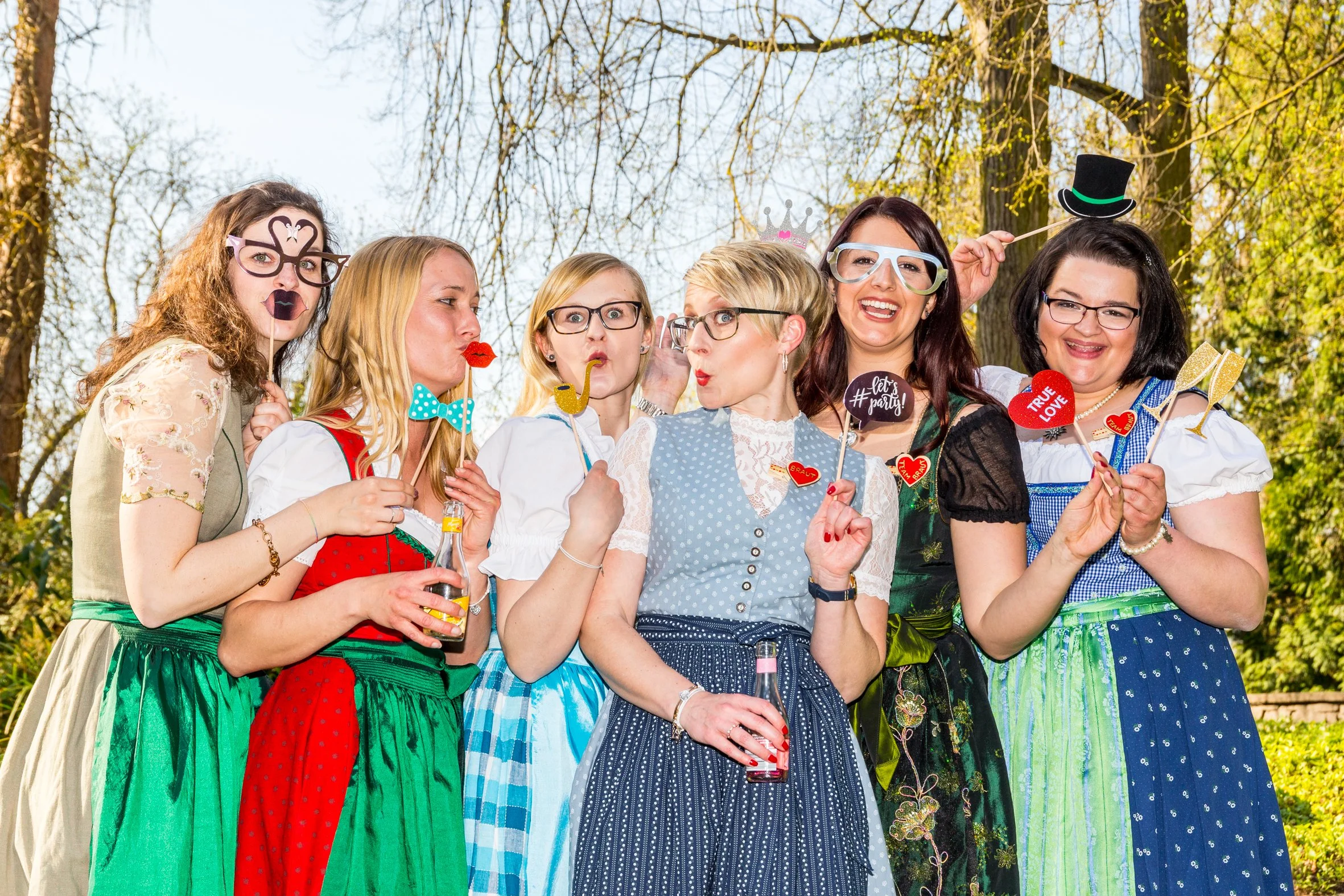 Eine Gruppe von sieben Frauen in traditionellen Trachten, die auf einer Wiese im Freien posieren, viele tragen lustige Accessoires und halten Fotoschilder mit positiven Botschaften, fröhliche Stimmung, Frühlings-/Sommerumgebung.