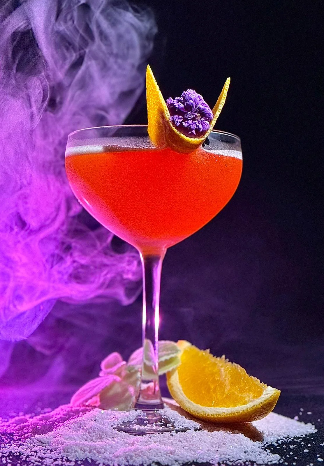 Ein roter Cocktail in einem stilvollen Glas mit einer Zitronenscheibe und einer Blume als Dekoration, mit blauem Rauch und Orangenscheiben im Hintergrund.