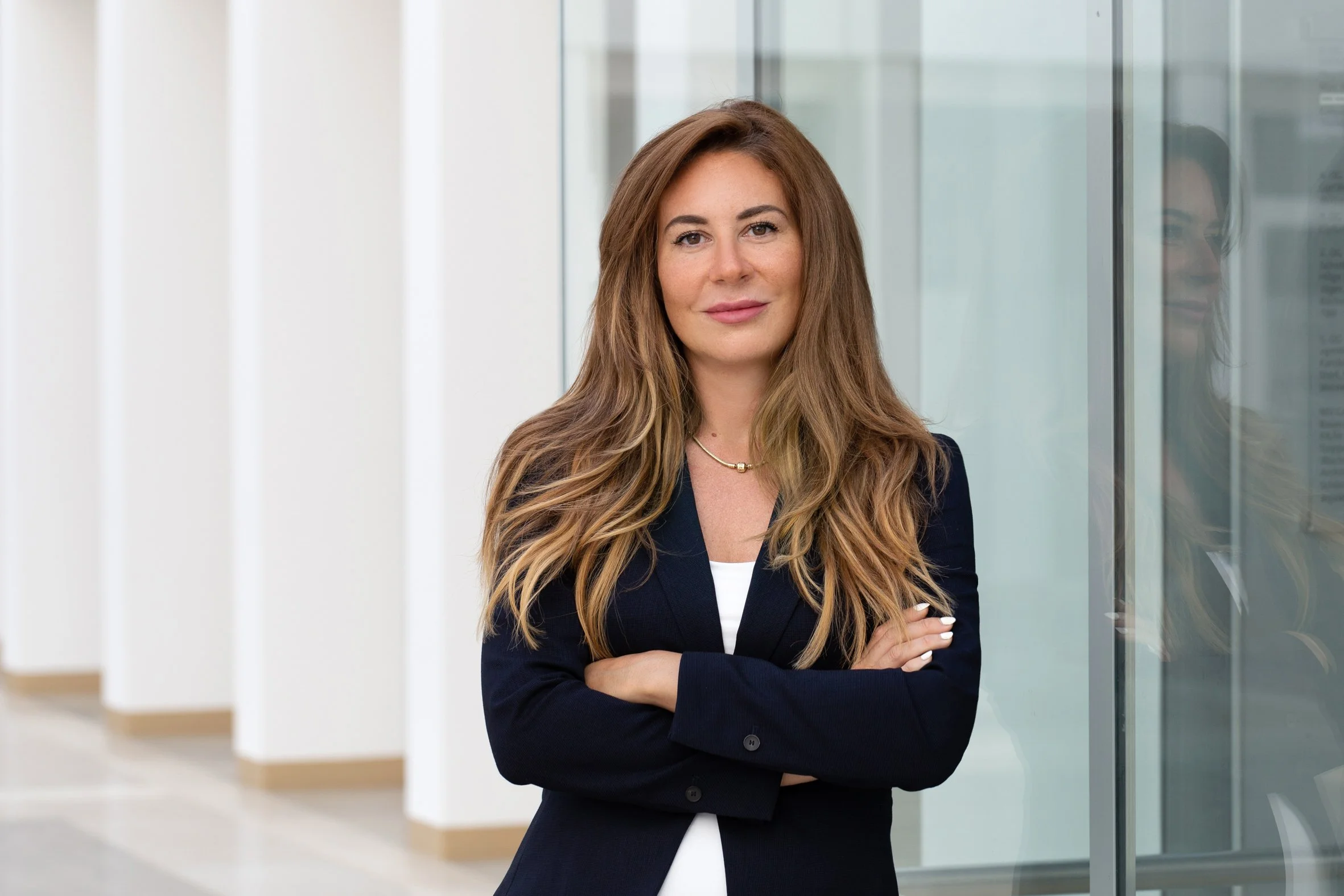 Eine Frau mit langem braunem Haar trägt einen dunklen Blazer und steht in einem modernen Bürogebäude, neben einer Glaswand mit reflektierendem Effekt.