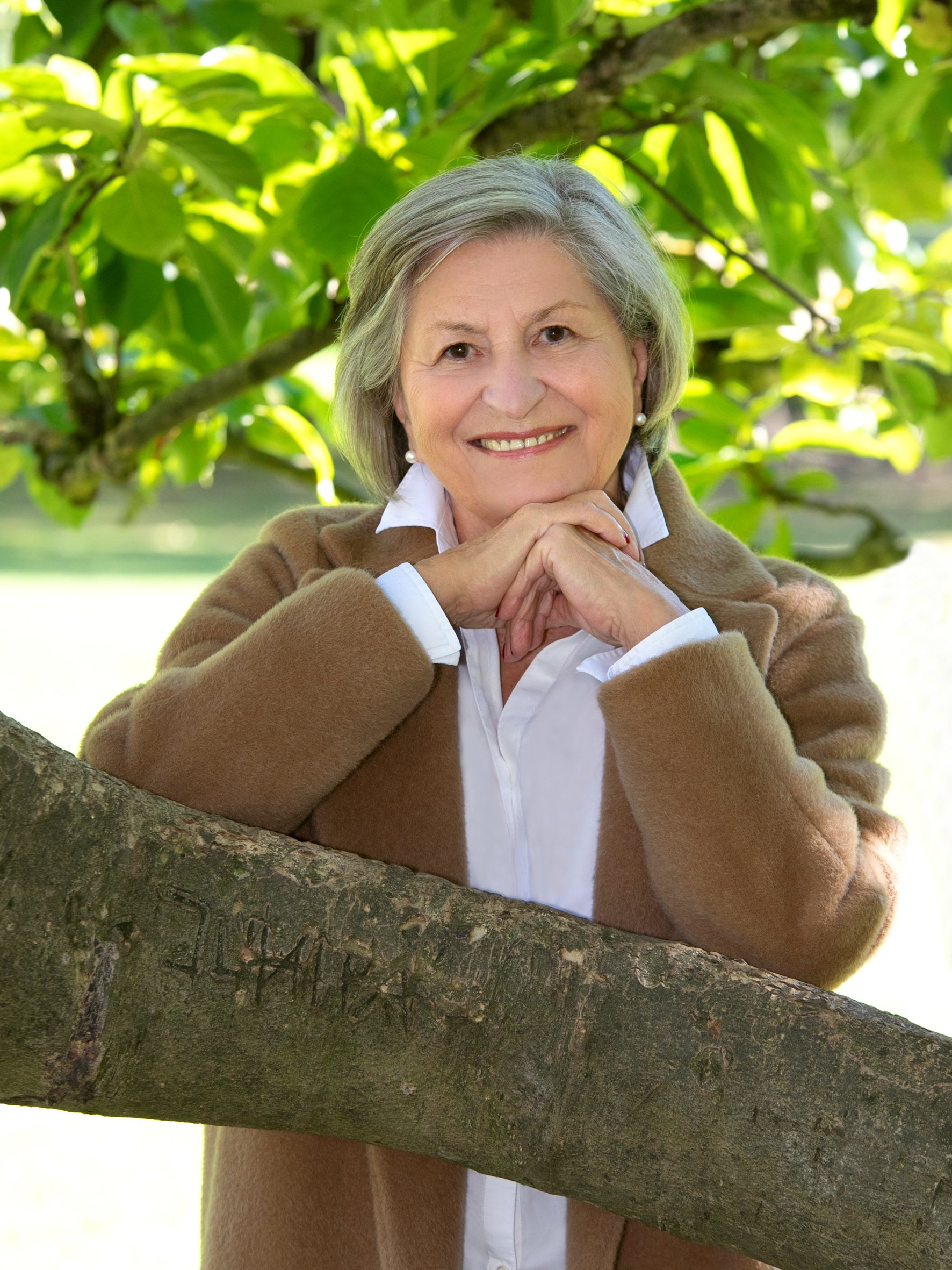 Alter Frau mit grauen Haaren, weißem Hemd, braunem Mantel, lächelnd, steht vor grünem Baum im Freien.