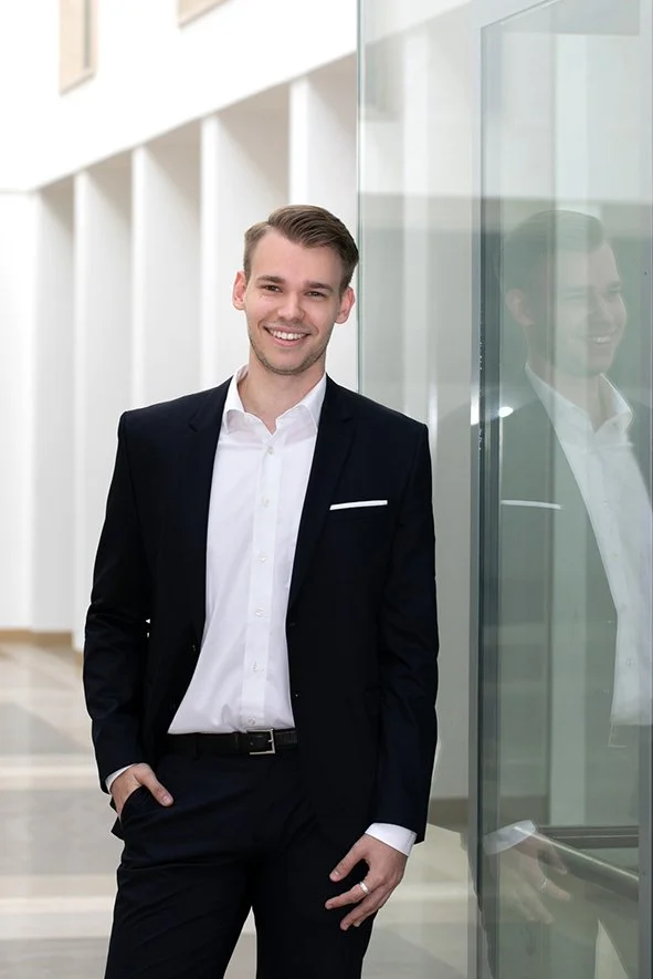 Ein junger Mann in einem schwarzen Anzug und weißem Hemd steht in einem modernen Gebäude und lächelt, während er die Hand in die Tasche steckt. Seine Reflexion ist in einer Glaswand neben ihm sichtbar.