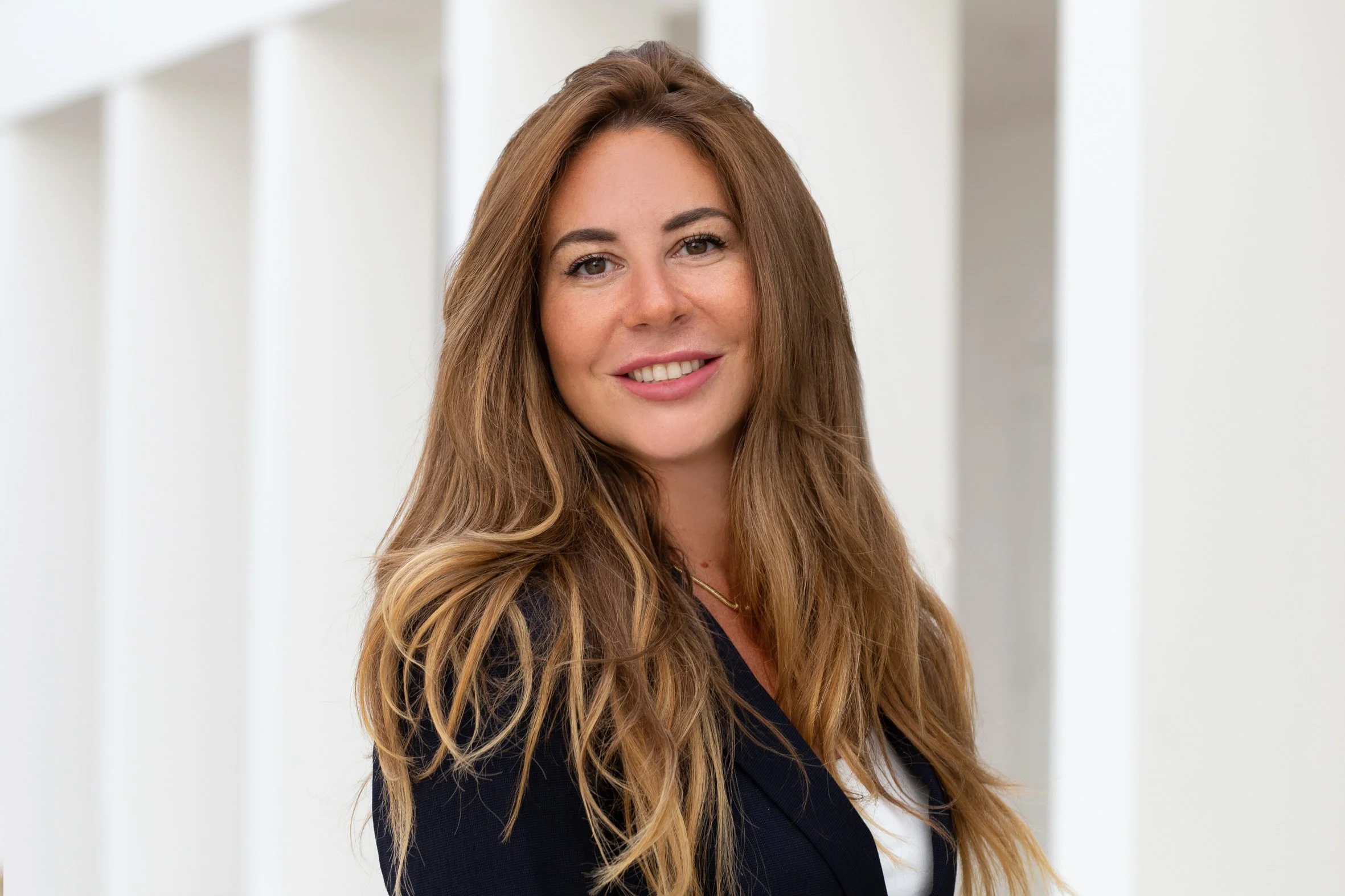 Eine junge Frau mit langen, lockigen hellbraunen Haaren, trägt eine schwarze Jacke und ein weißes Top, steht vor einem hellen, modernen Hintergrund.