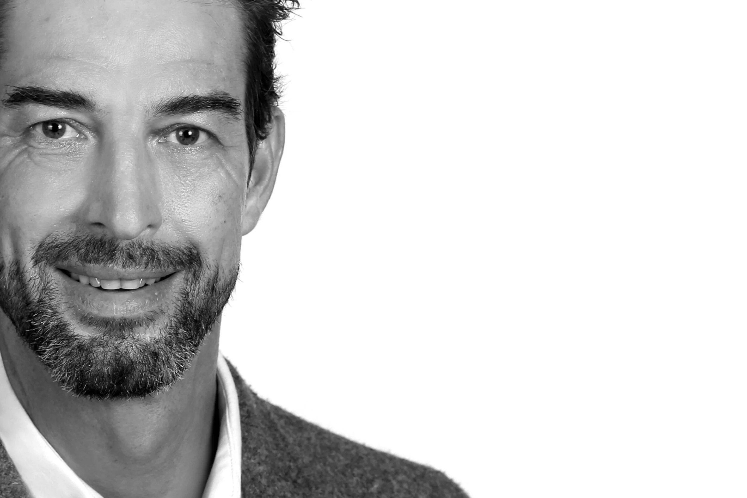 Nahaufnahme eines lächelnden Mannes mit dunklem Haar und Bart, in schwarz-weiß.
