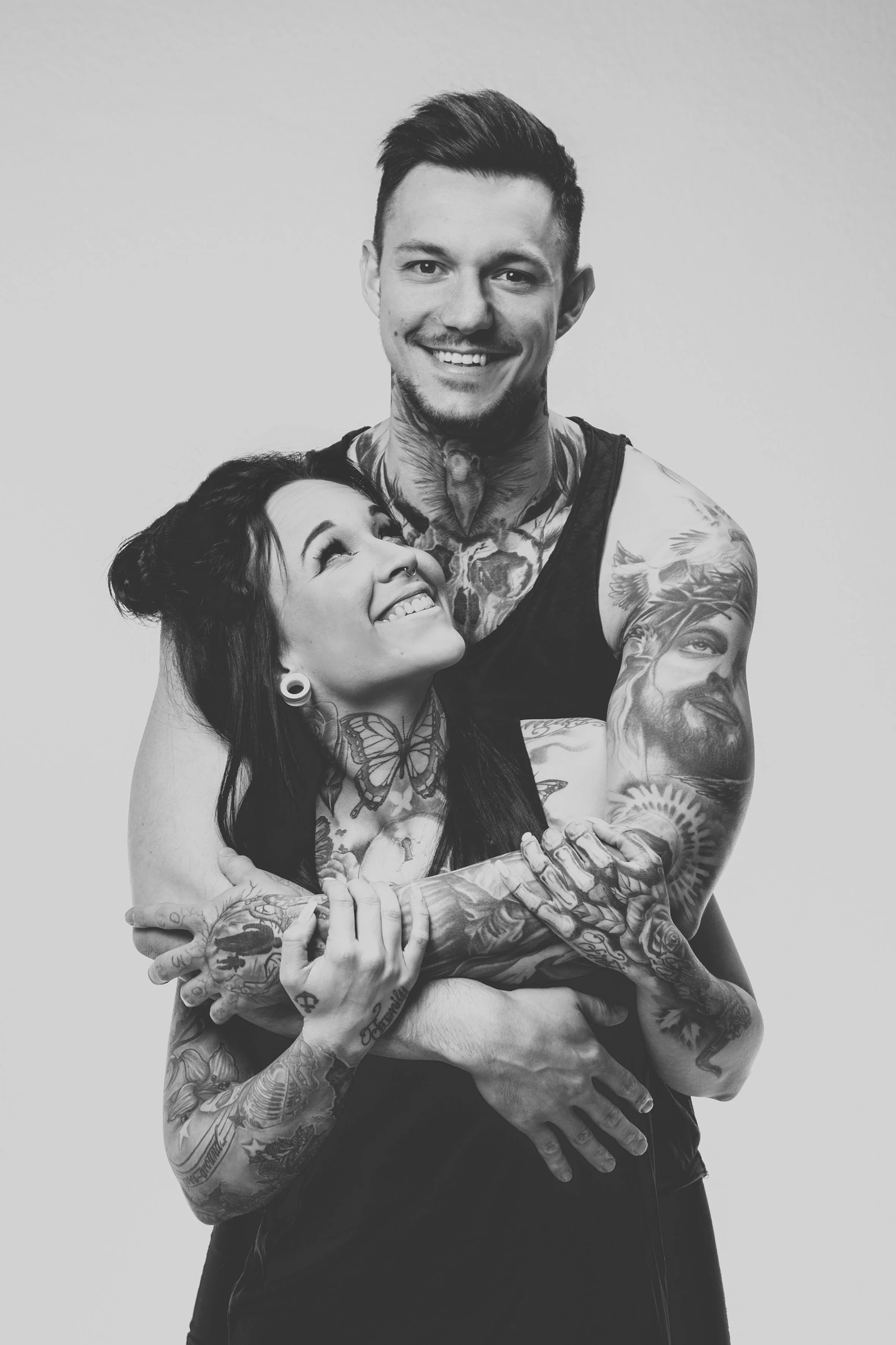 Ein lachendes Paar mit Tattoos umarmt sich gegen einen hellen Hintergrund.
