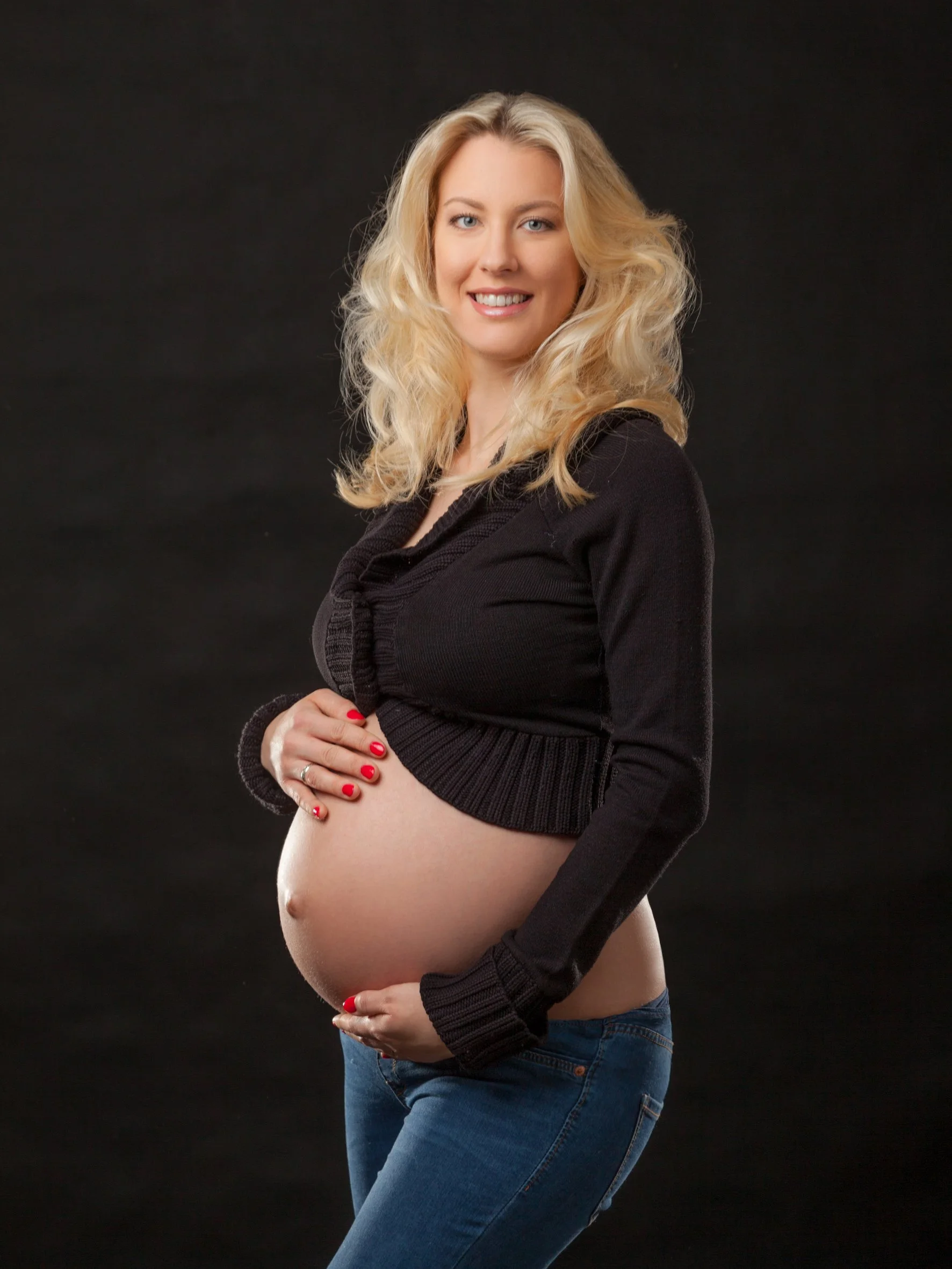 Schwangere Frau mit langer Blondhaar, die ein schwarzes Oberteil und Jeans trägt, hält ihre Babybauch mit beiden Händen, vor dunklem Hintergrund.