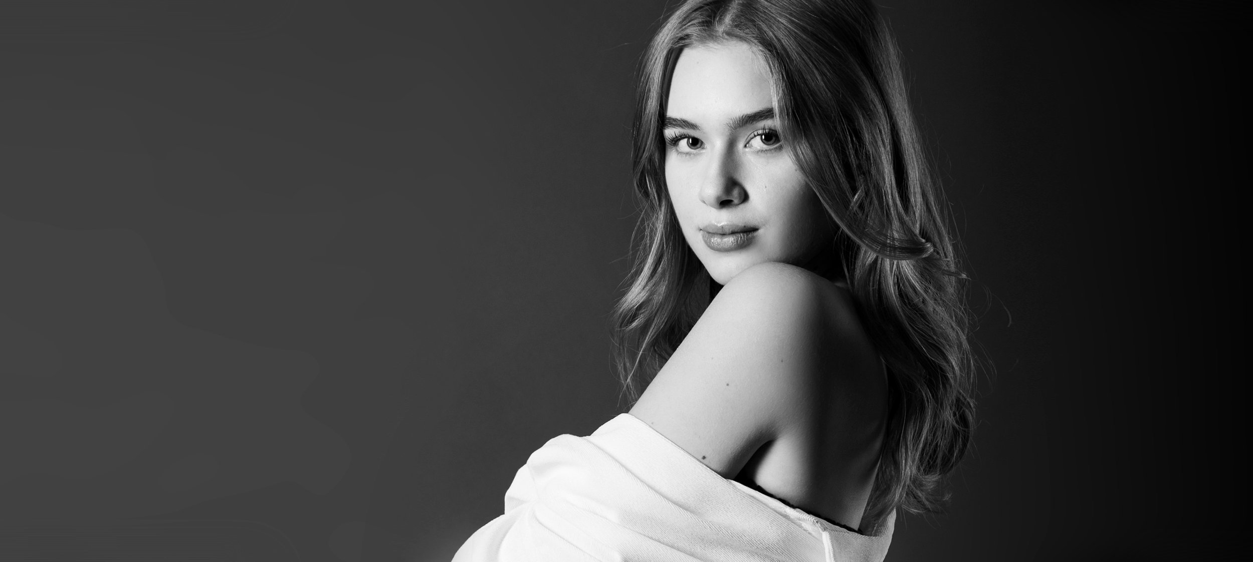 Schwarz-weiß-Fotografie eines jungen Mädchens mit lockigem Haar, halb blickt sie in die Kamera, trägt ein weißes Oberteil, vor dunklem Hintergrund.