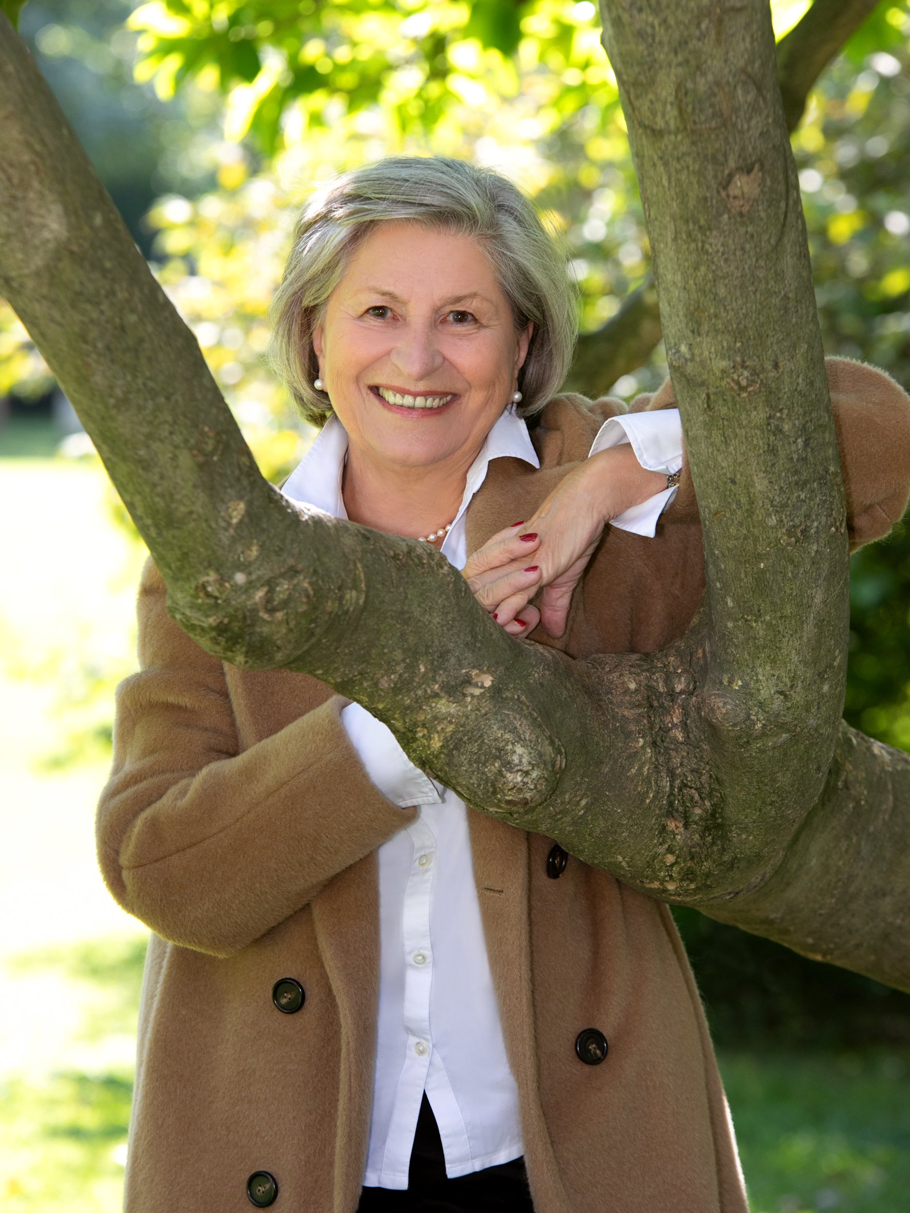 Eine ältere Frau steht hinter einem Baum und lächelt in die Kamera, während sie die Arme um den Baum legt.