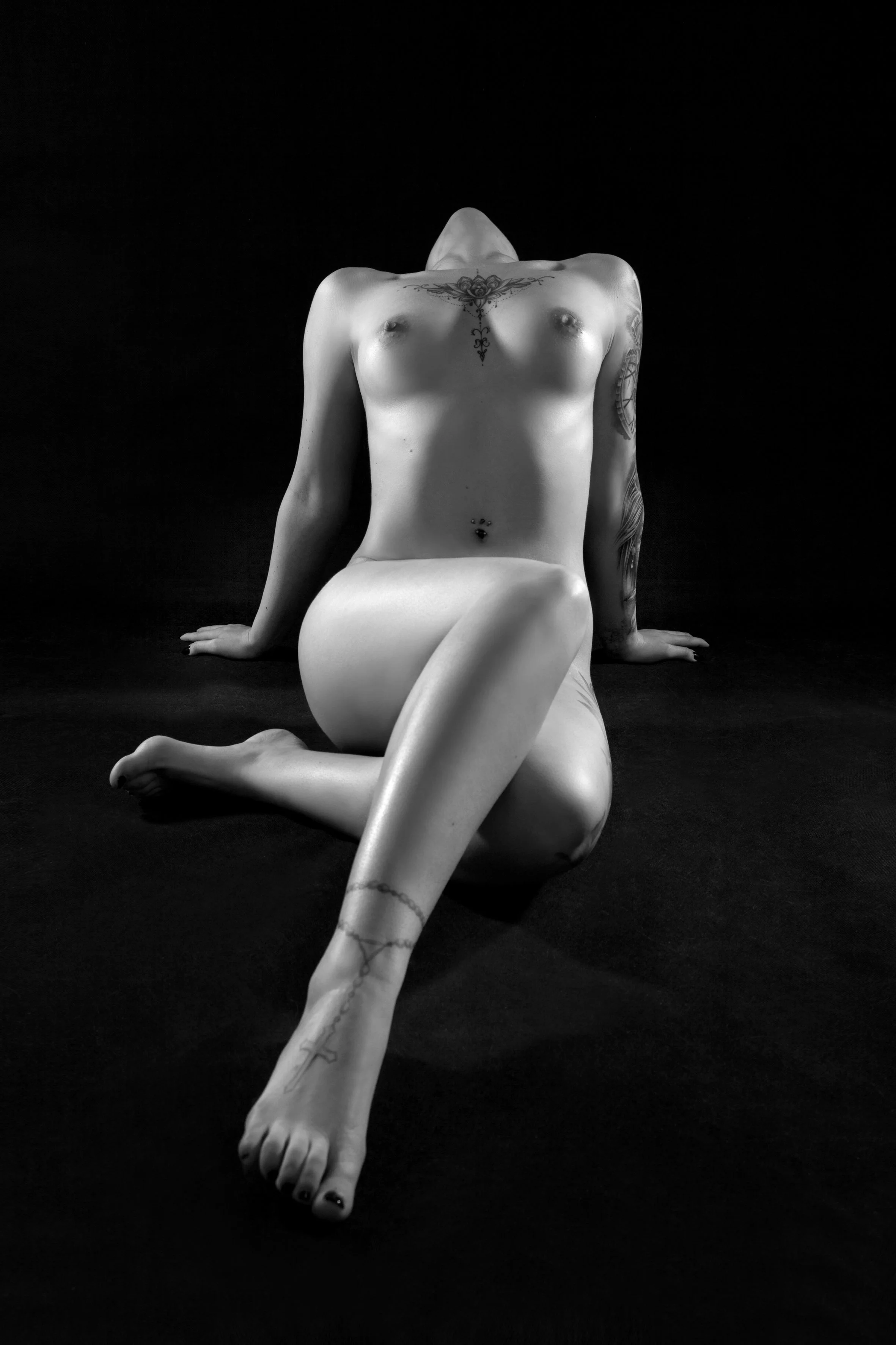 Schwarz-weiß-Foto einer nackten Frau mit Tattoos, die auf dem Boden sitzt, die Beine gekreuzt, Kopf nach hinten geneigt, auf einem schwarzen Hintergrund.