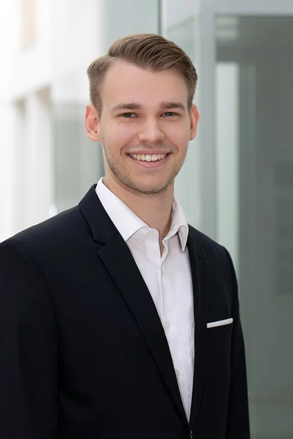 Ein junger Mann in formeller Kleidung, der vor einem modernen Bürogebäude lächelt.