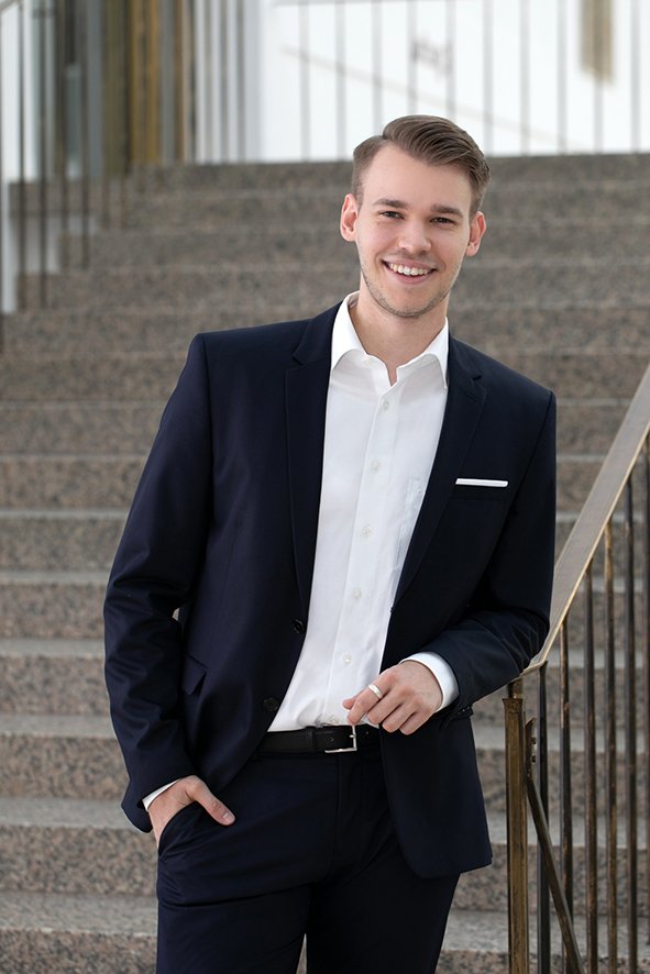 Junger Mann in dunklem Anzug und weißem Hemd steht auf einer Treppe und lächelt.