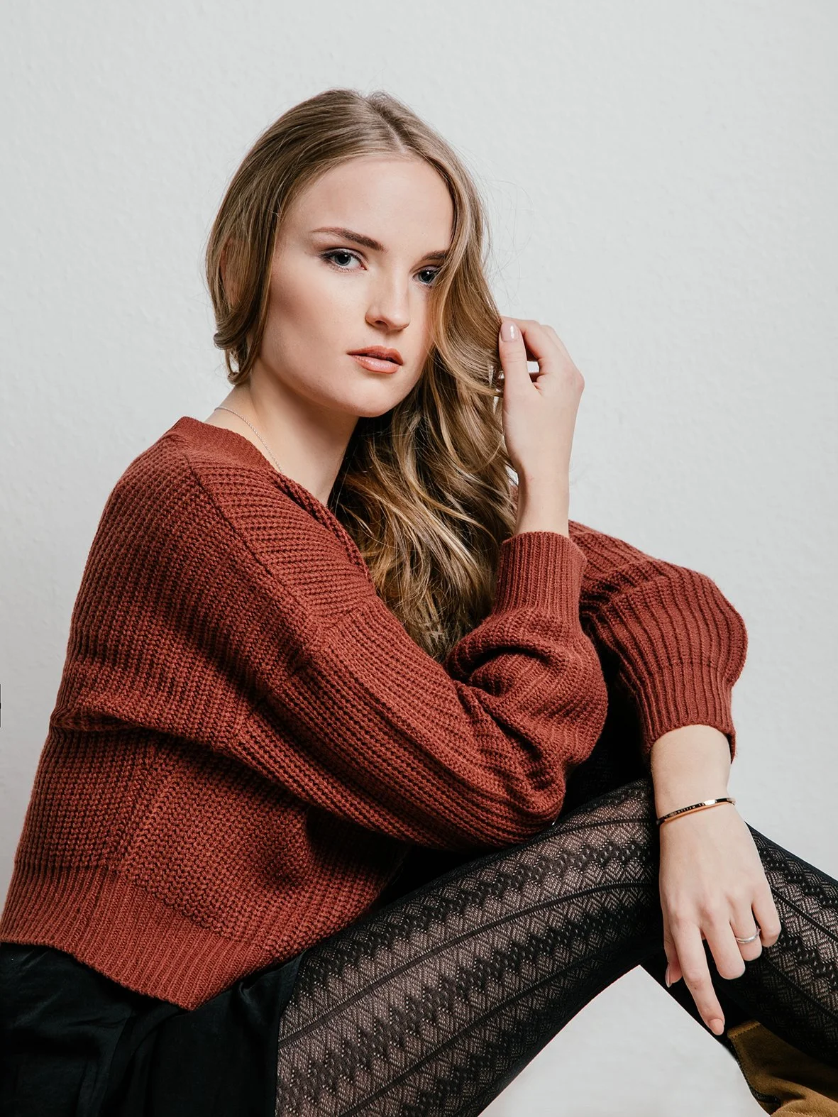 Junge Frau mit langem, lockigem Haar, trägt einen maronenfarbenen Pullover und schwarze, gemusterte Leggings, sitzt vor weißer Wand.