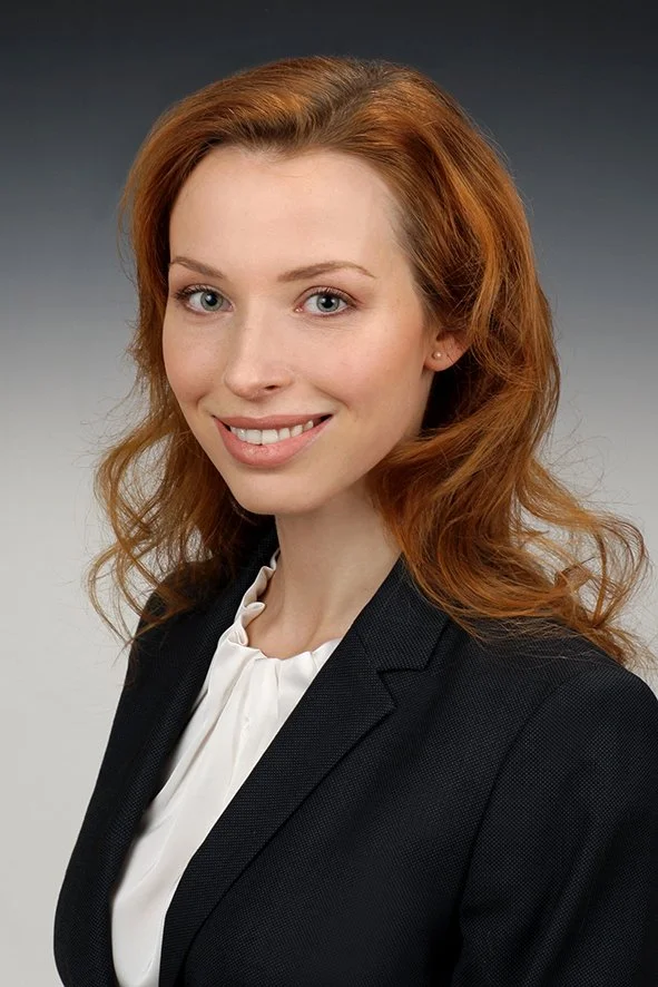 Porträt einer jungen Frau mit roten Haaren in formeller Kleidung vor einem grauen Farbverlauf Hintergrund.