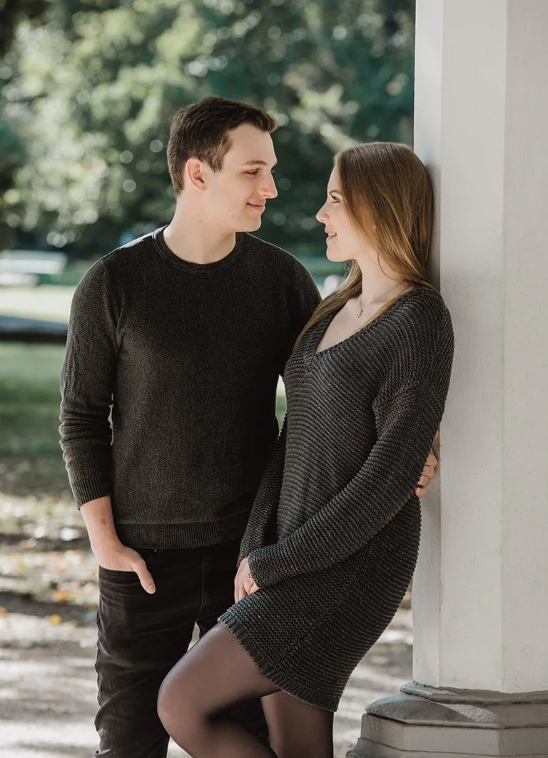 Ein Mann und eine Frau stehen sich gegenüber, lächeln und schauen sich verliebt an, während die Frau an einer Säule lehnt.