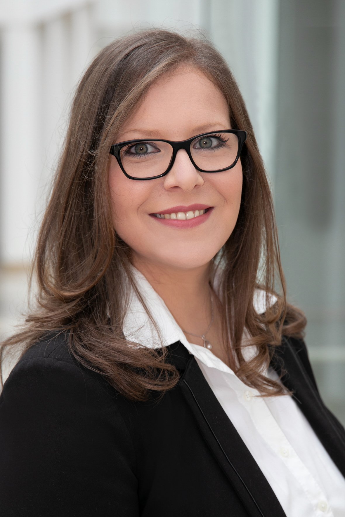 Porträt einer lächelnden Frau mit Brille in formellem Business-Outfit vor einem modernen Hintergrund.