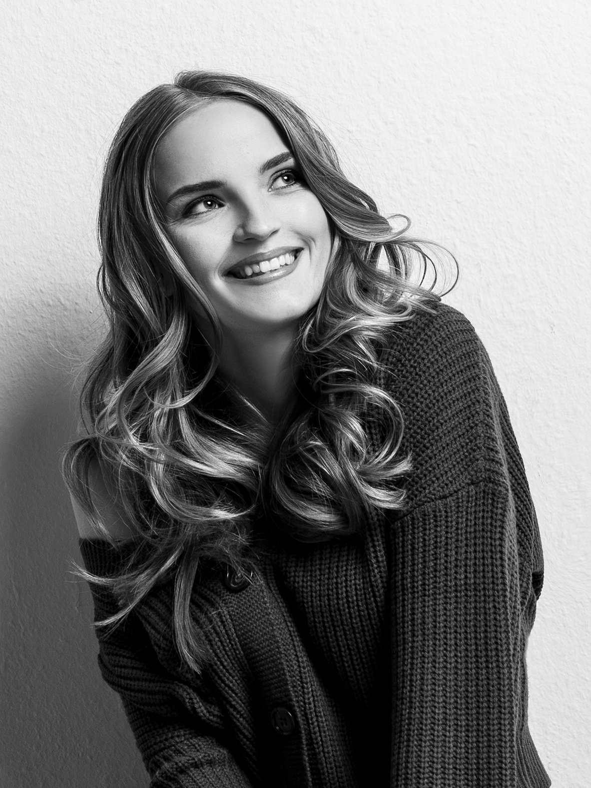 Schwarze und weiße Porträtaufnahme einer lachenden jungen Frau mit gewellten Haaren, die eine dunkle Strickjacke trägt, vor einer hellen Wand.