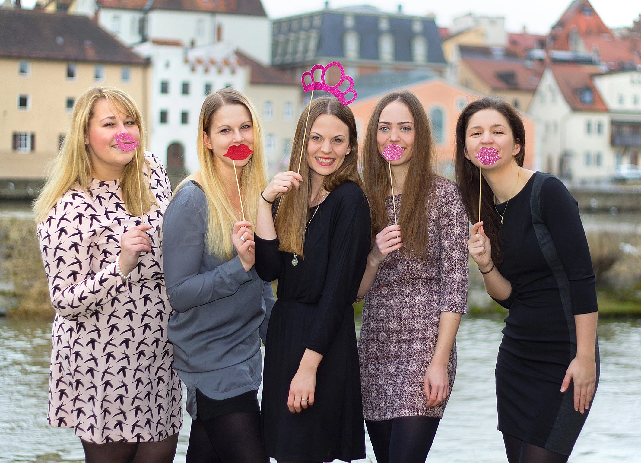 Fünf Frauen stehen am Flussufer, halten Piercings aus glitzerndem Papier auf Spiesse und posieren für ein Foto, im Hintergrund bunte Häuser in einer Stadt.
