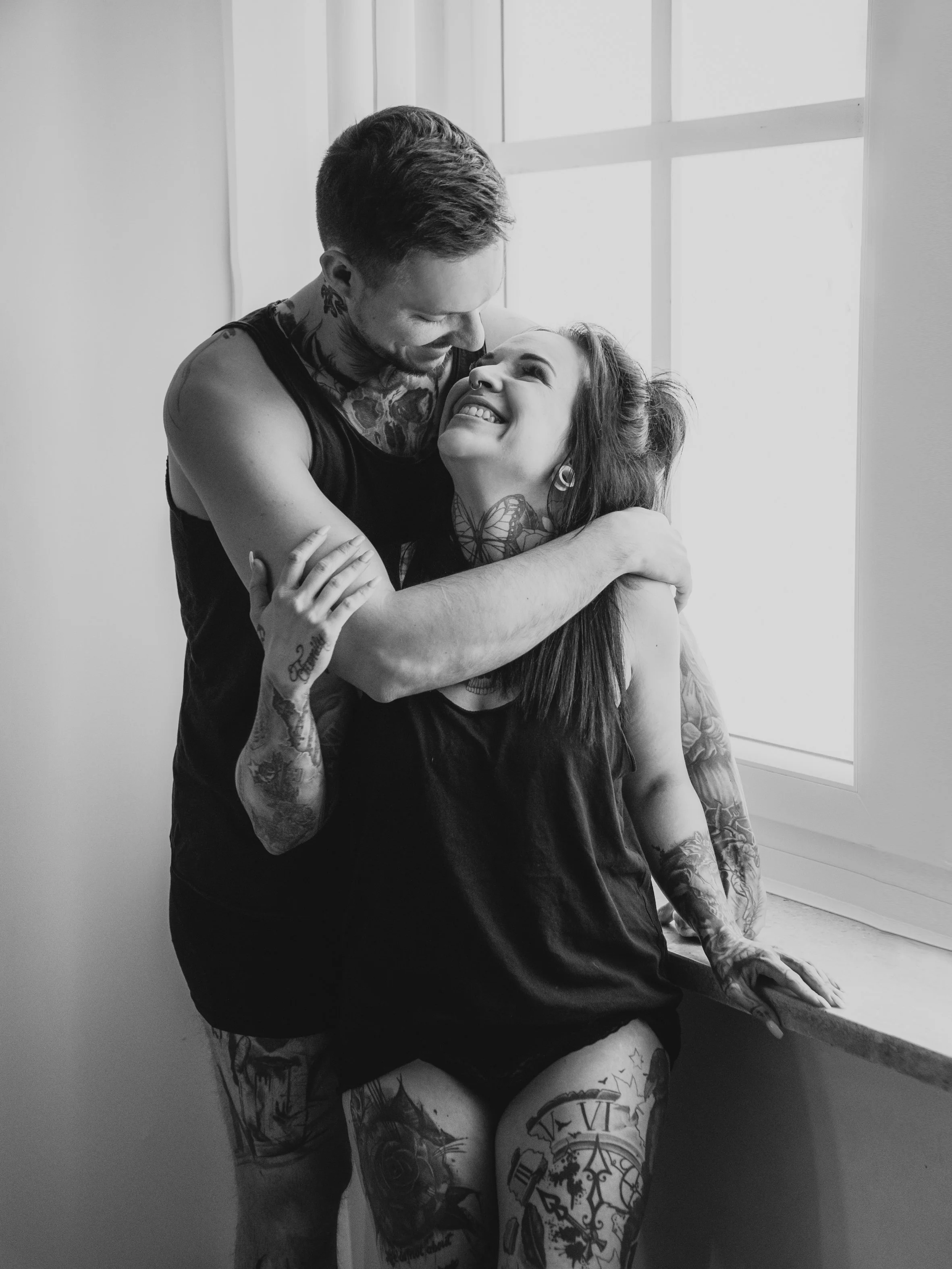 Blick auf ein lachendes Paar, das sich umarmt, im Raum vor einem Fenster, Schwarz-Weiß-Fotografie.