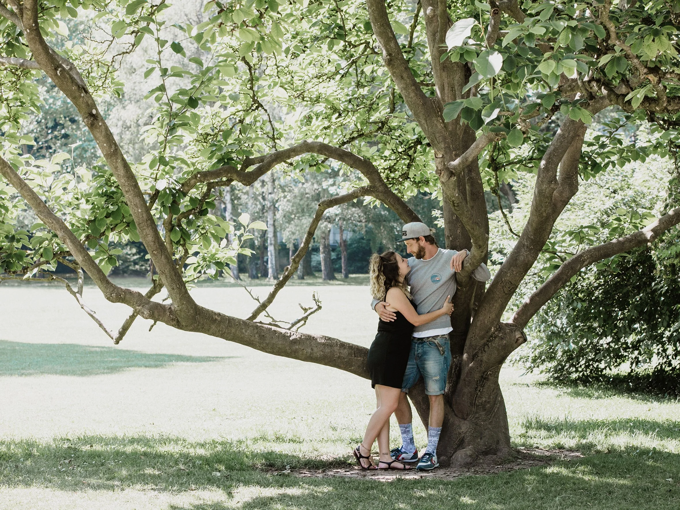 Ein Paar steht eng an einem Baum im Park, lächelt sich liebevoll an.