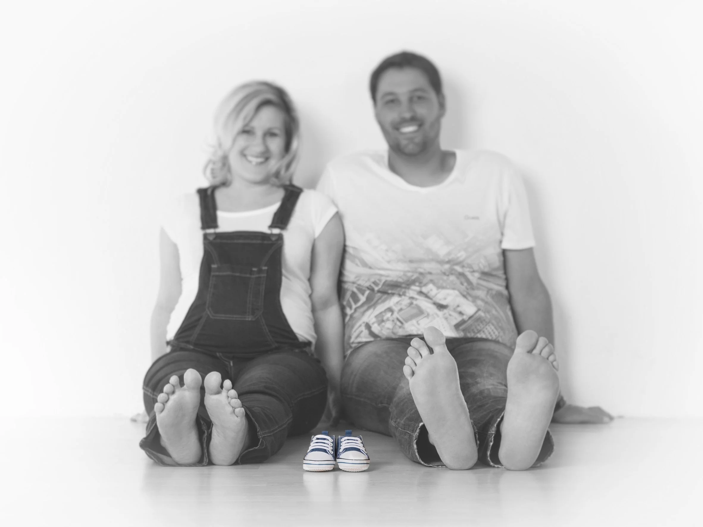 Ein lachendes Paar sitzt barfuß auf dem Boden, die Frau trägt Overalls und der Mann ein T-Shirt, zwischen ihnen ein Paar kleine Kinderschuhe.