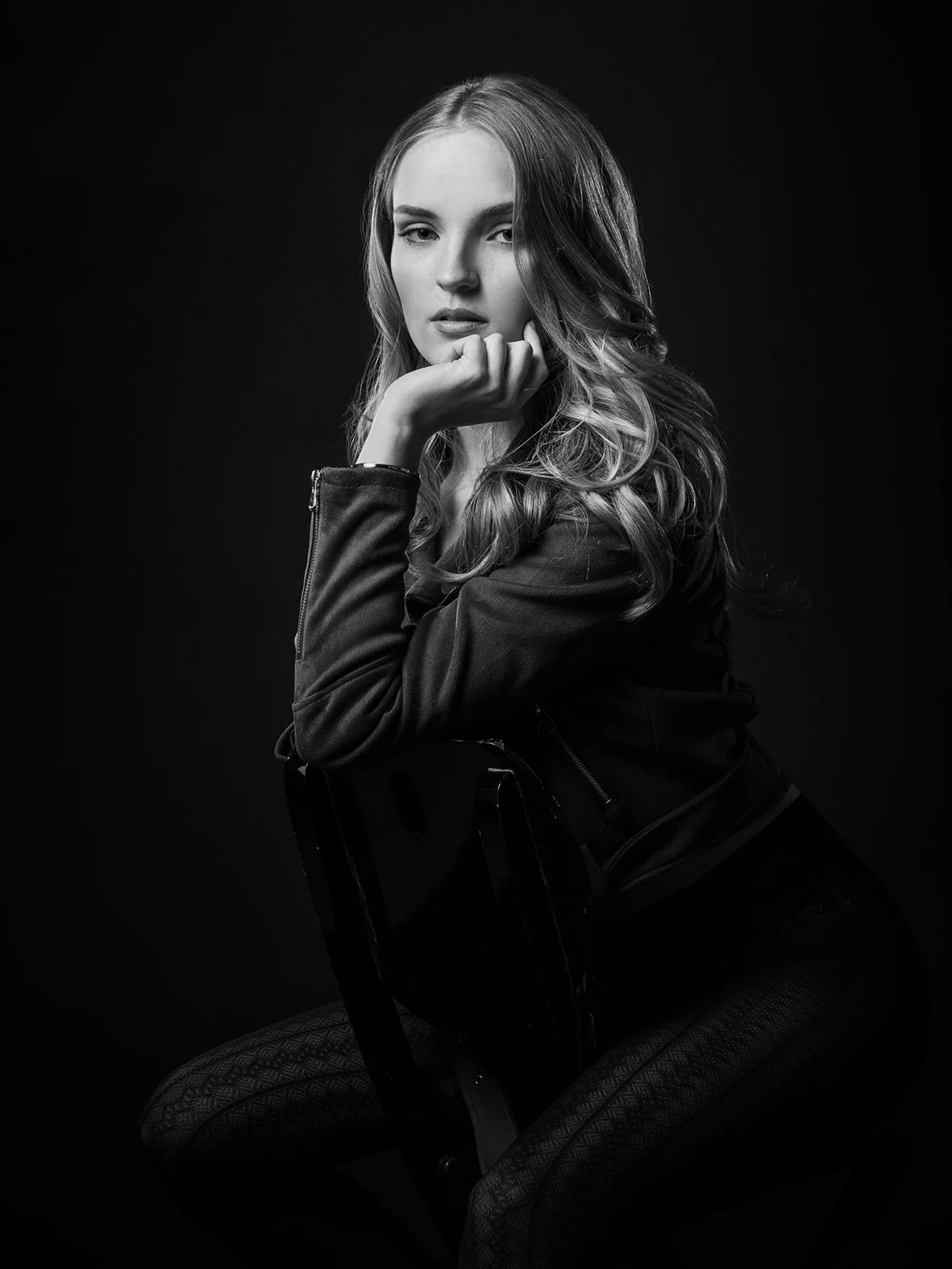 Schwarz-weiß Porträt einer jungen Frau, die auf einem Stuhl sitzt, mit langem, lockigem Haar, blickt aufmerksam, trägt eine Lederjacke, dunkler Hintergrund.