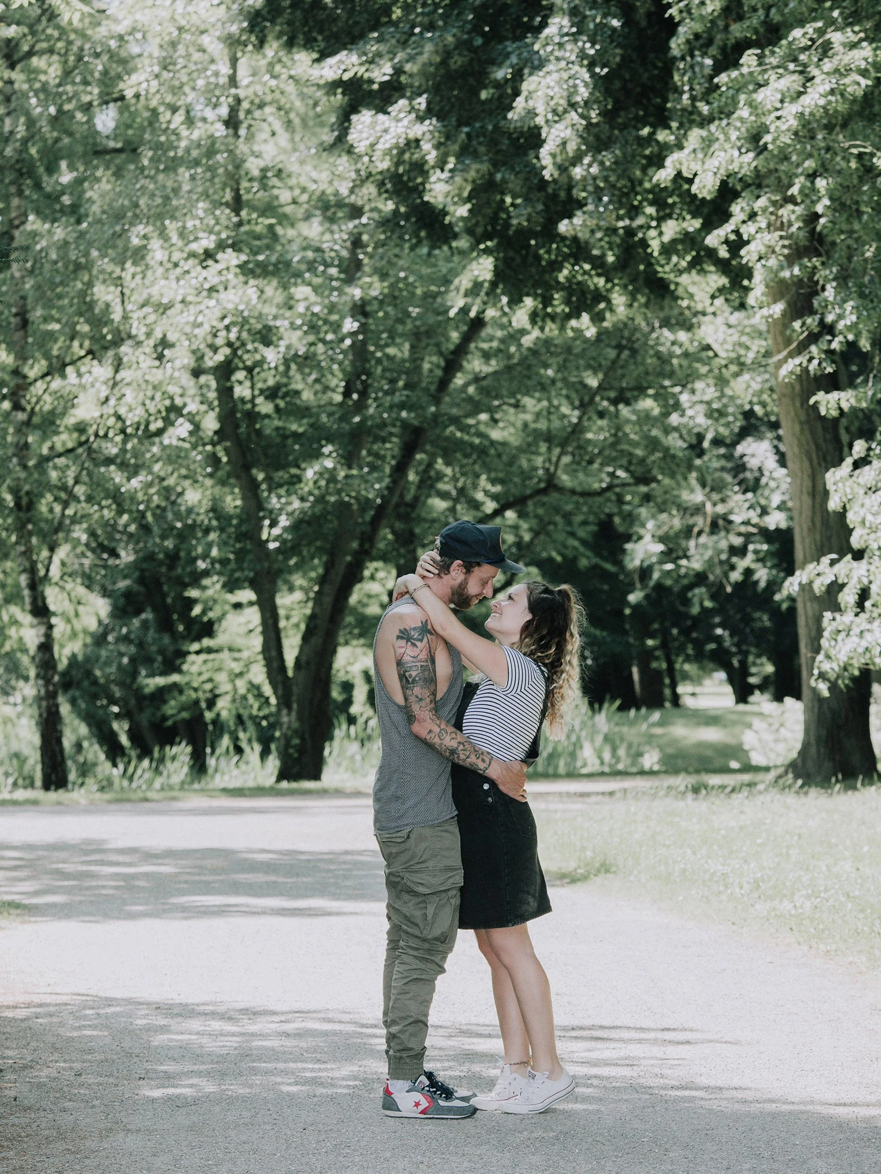 Ein junges Paar umarmt sich im Park. Er trägt eine Mütze und hat Tattoos, sie trägt ein gestreiftes T-Shirt und einen schwarzen Rock. Im Hintergrund sind grüne Bäume und ein sonniger Tag.
