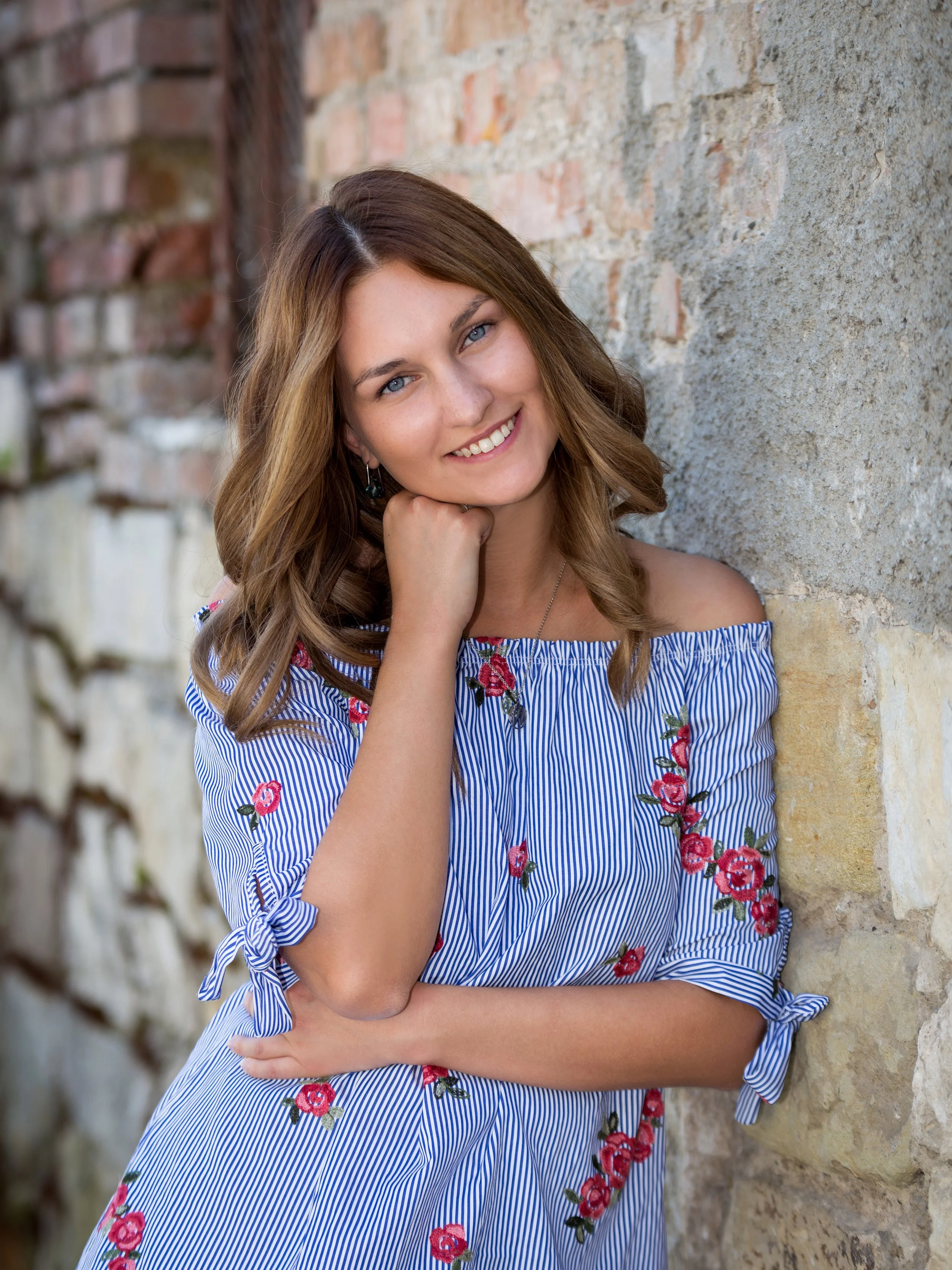 Lächelnde junge Frau mit braunen Locken, die in einem blauen, gestreiften Kleid mit roten Blumenmuster steht neben einer Steinaufmauer, die auf der linken Seite im Bild sichtbar ist.