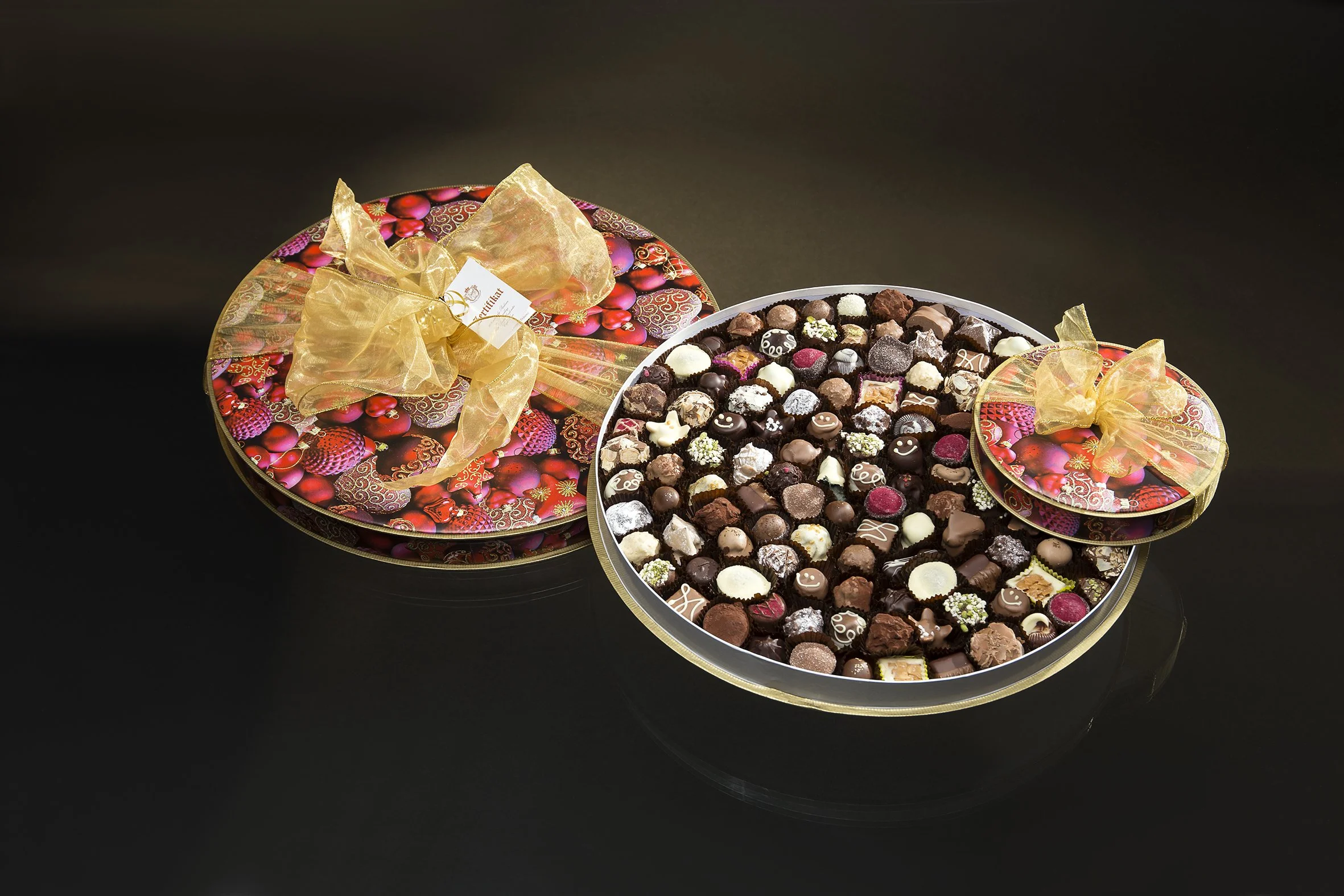 Ein runder Geschenkkarton mit bunter Schokoladensammlung, verziert mit goldenen Schleifen, auf schwarzem Hintergrund.
