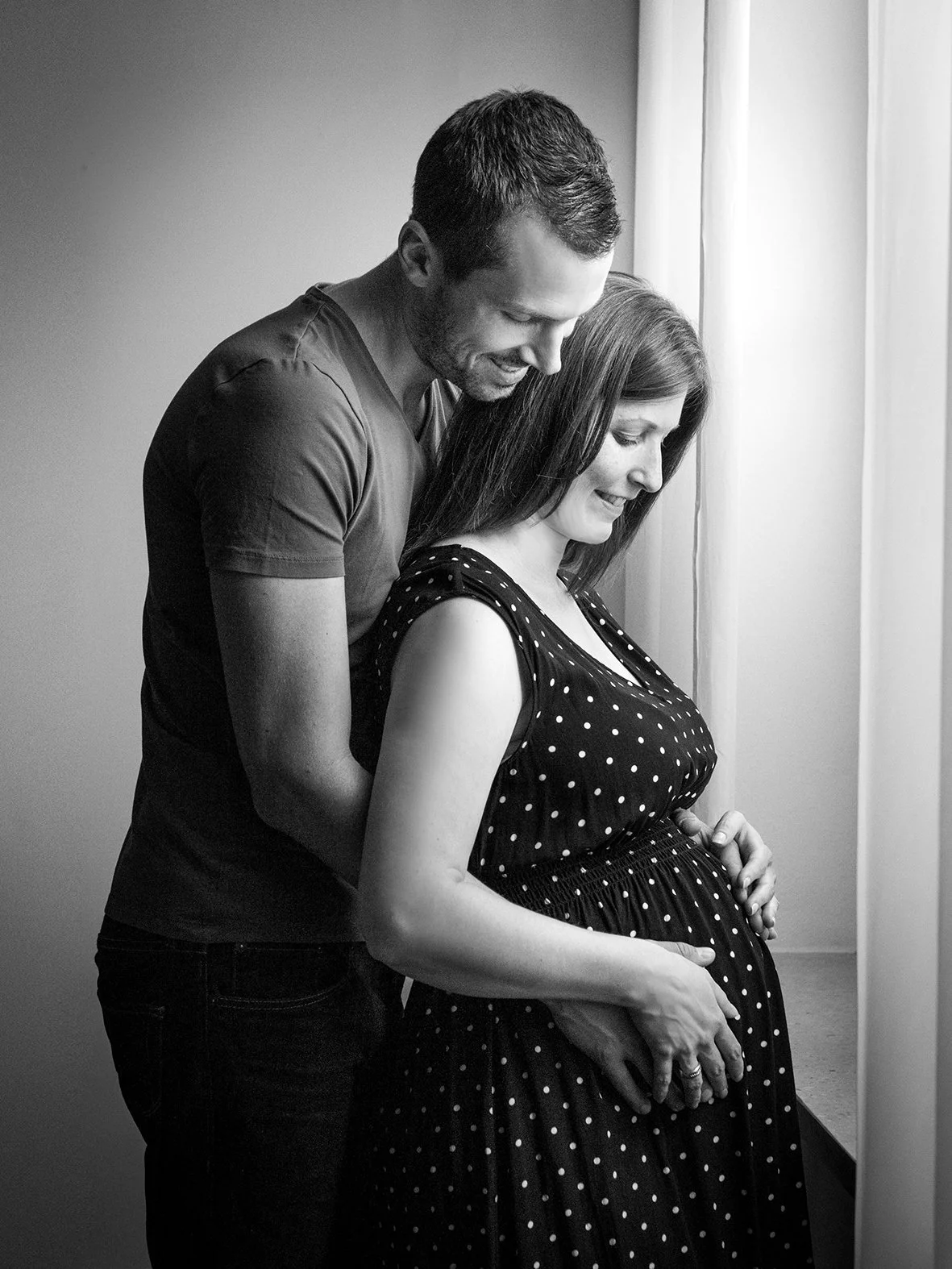 Schwangere Frau mit langem Haar, die von einem lächelnden Mann umarmt wird, beide schauen liebevoll auf den Bauch, in einem Raum neben einem Fenster mit Vorhang, schwarz-weiß Foto.