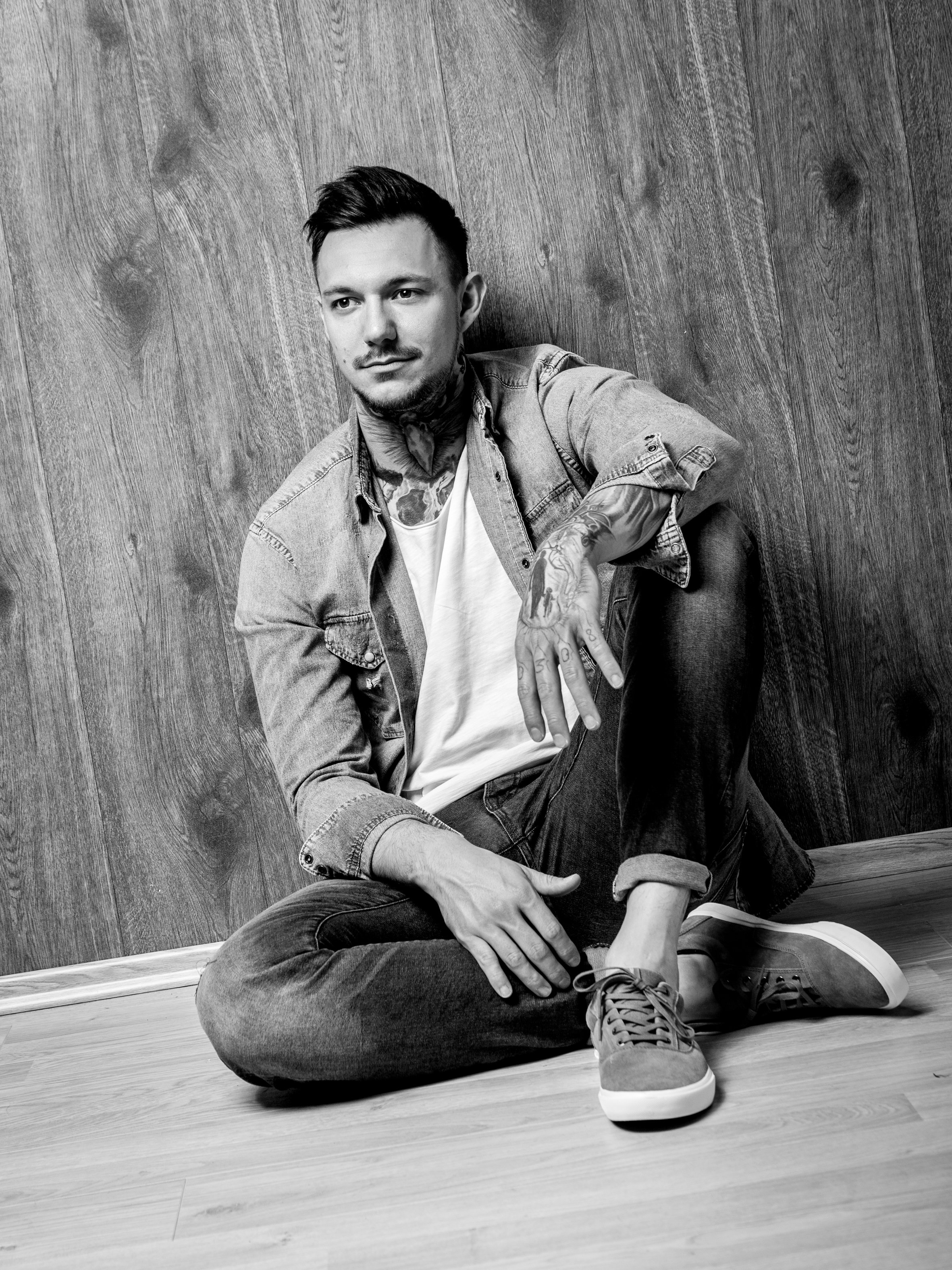 Schwarz-Weiß-Foto eines jungen Mannes, der auf dem Boden sitzt und gegen eine Holzwand lehnt, trägt Jeansjacke, T-Shirt, Sneakers und hat Tätowierungen an Armen und Hals.