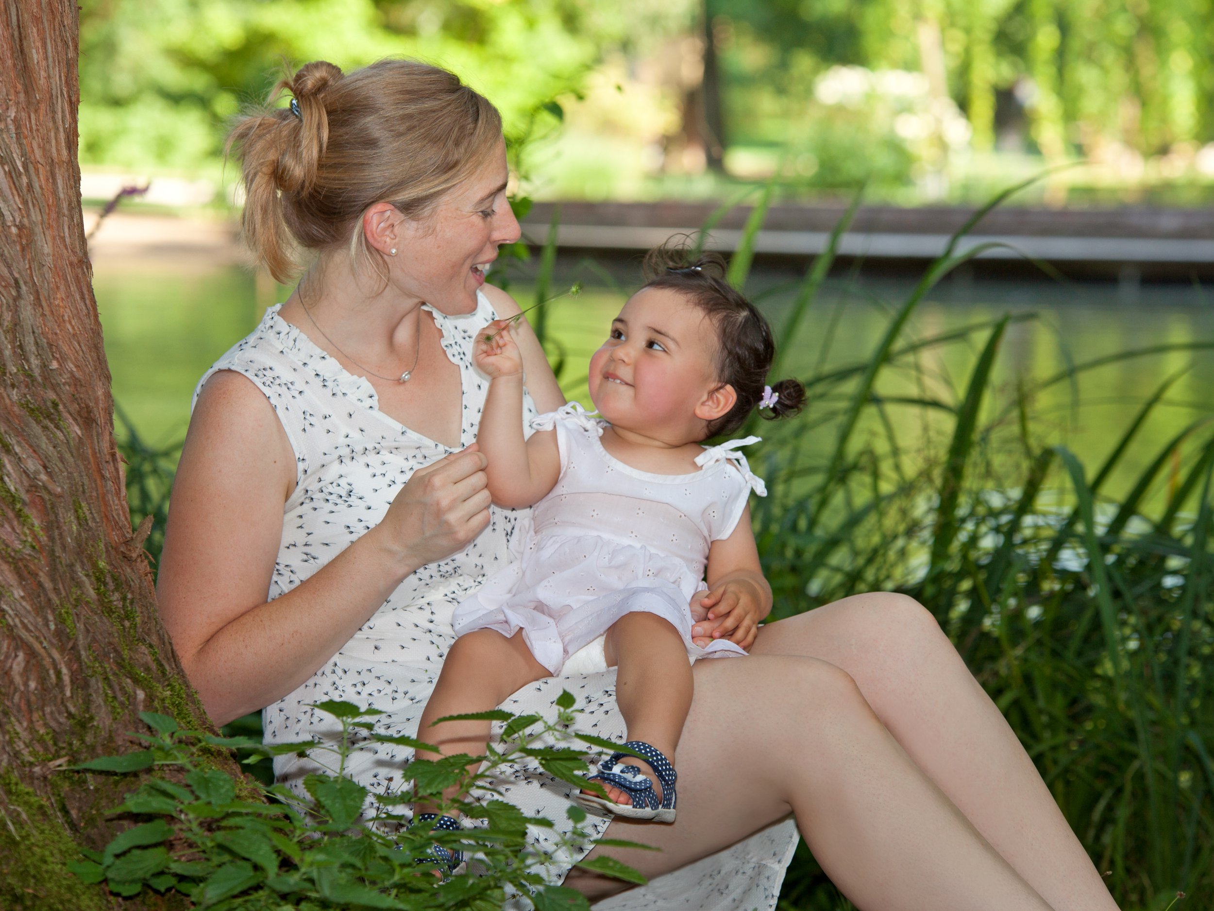 Eine Frau und ein kleines Mädchen sitzen am Ufer eines Flusses, umgeben von grünen Pflanzen und Bäumen, im Freien bei sonnigem Wetter.