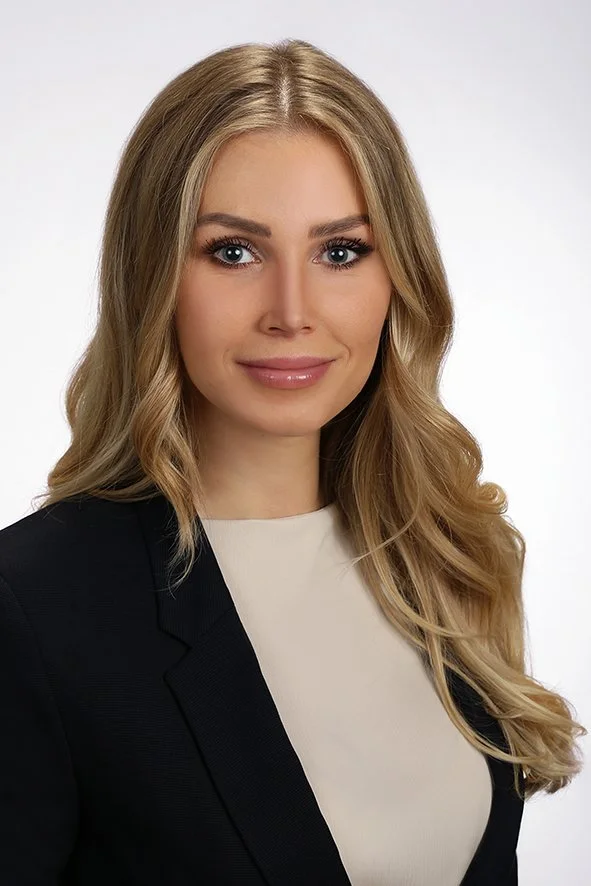 Porträt einer jungen Frau mit langen, gewellten blonden Haaren, blauen Augen, weißem Hemd und schwarzer Blazer vor neutralem Hintergrund.