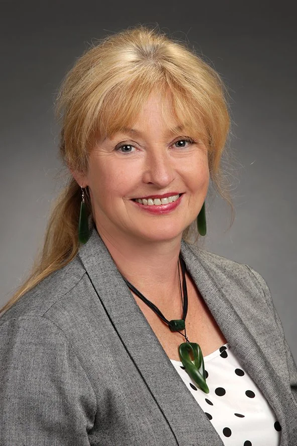 Ein portraitierte Frau mit blonden Haaren, grauem Blazer, einem weißen Top mit schwarzen Punkten, grünen Ohrringen und einer grünen Halskette vor grauem Hintergrund.