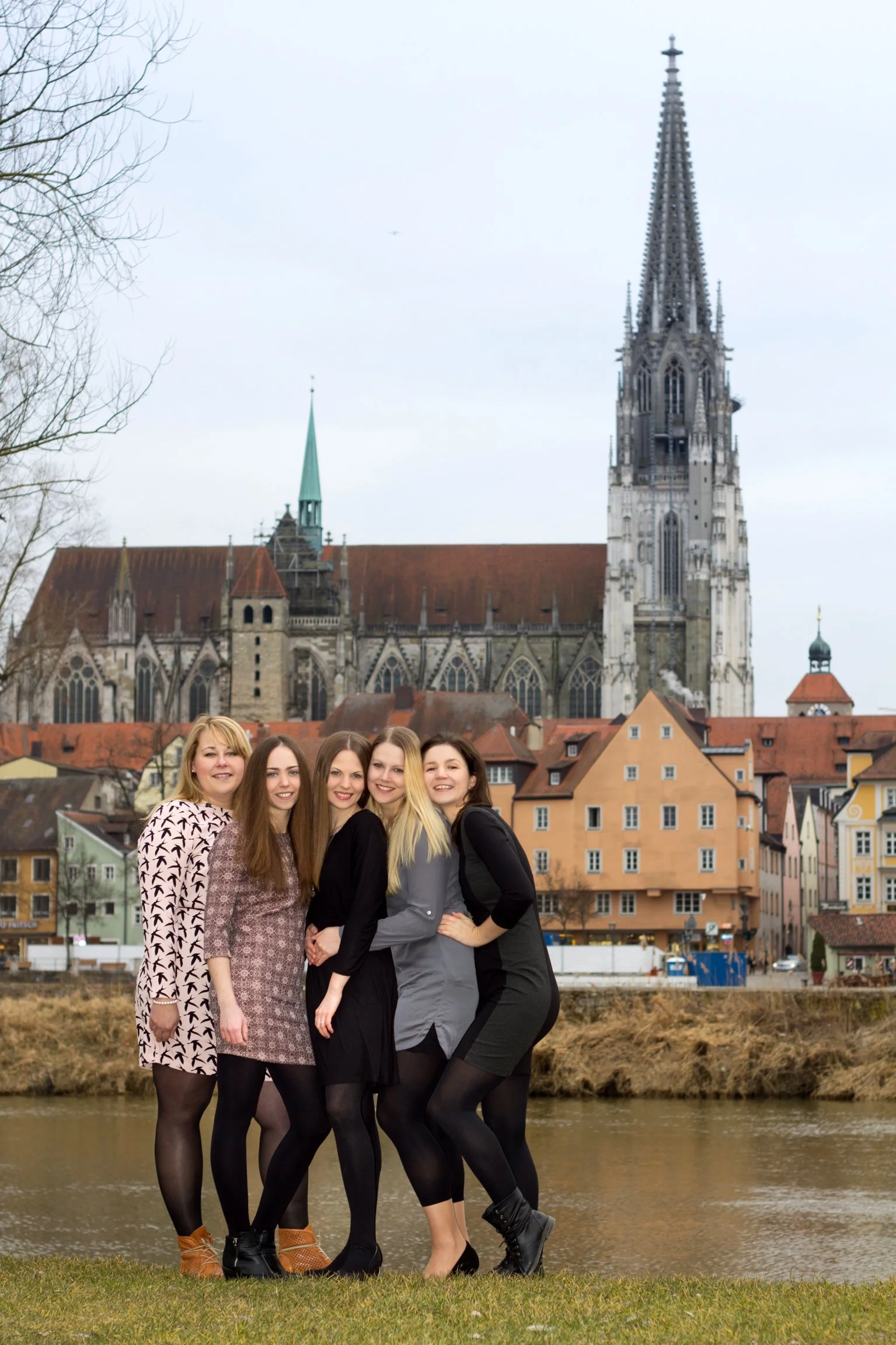 Fünf Frauen stehen am Flussufer vor einer Stadt mit bunten Häusern und einer gotischen Kirche im Hintergrund.